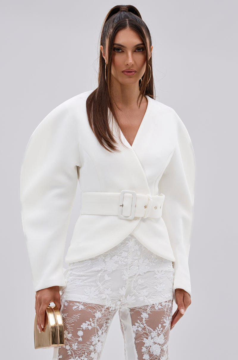 A woman wears the LANA SCUBA BLOUSE, featuring sculptural white sleeves and a cinched waist with a wide buckle belt, paired with sheer lace pants. She holds a gold clutch and her long hair is styled in a high ponytail against a plain light background.