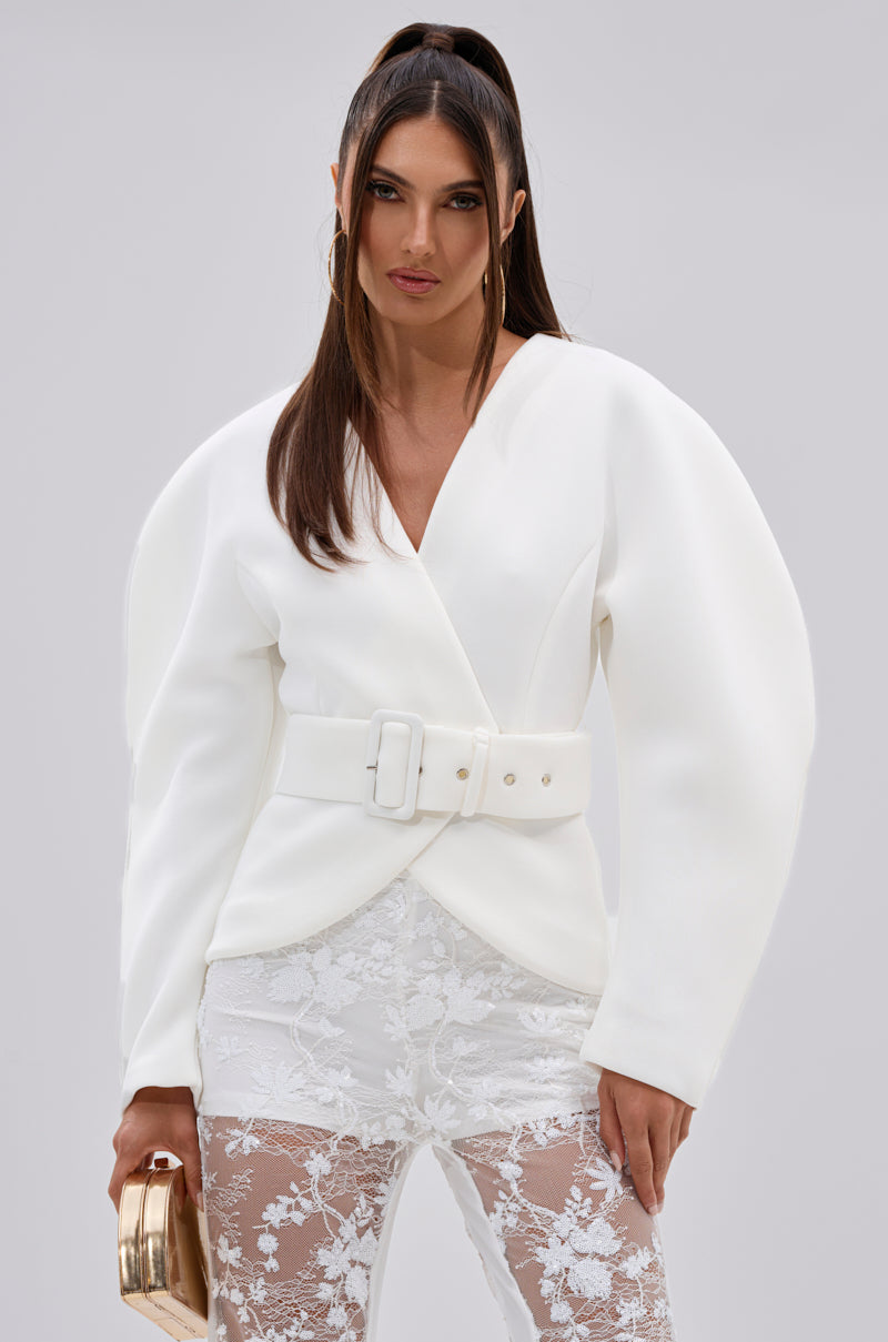 A woman with long brown hair models the LANA SCUBA BLOUSE, a stylish white belted piece with sculptural sleeves, paired with sheer lace pants and a gold clutch, posing against a plain light gray background.