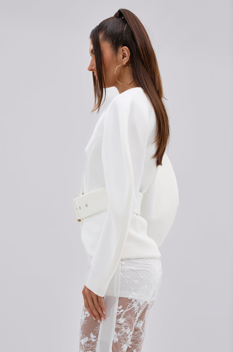 A woman stands in profile against a light gray background, wearing the LANA SCUBA BLOUSE with sculptural sleeves and a cinched waist, paired with sheer lace pants. She has long brown hair in a high ponytail and large hoop earrings.