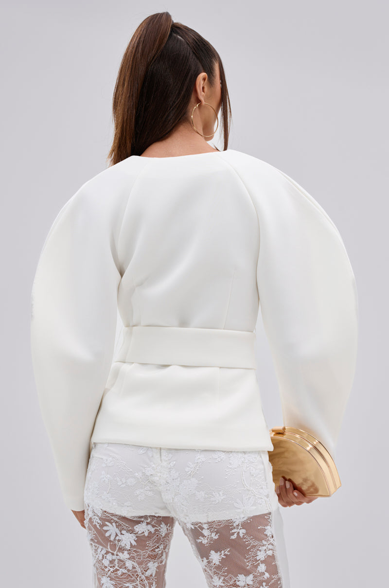 Woman with long brown hair in a ponytail is seen from behind wearing the LANA SCUBA BLOUSE, styled with white lace pants, large hoop earrings, and a small beige clutch against a plain background.