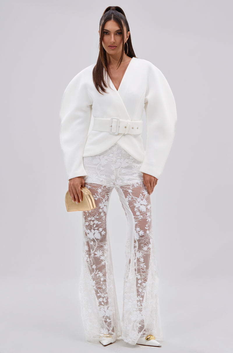 A woman poses in the LANA SCUBA BLOUSE, featuring a white belted design with sculptural sleeves, paired with sheer white lace pants, pointed-toe heels, and a gold clutch against a plain light background.