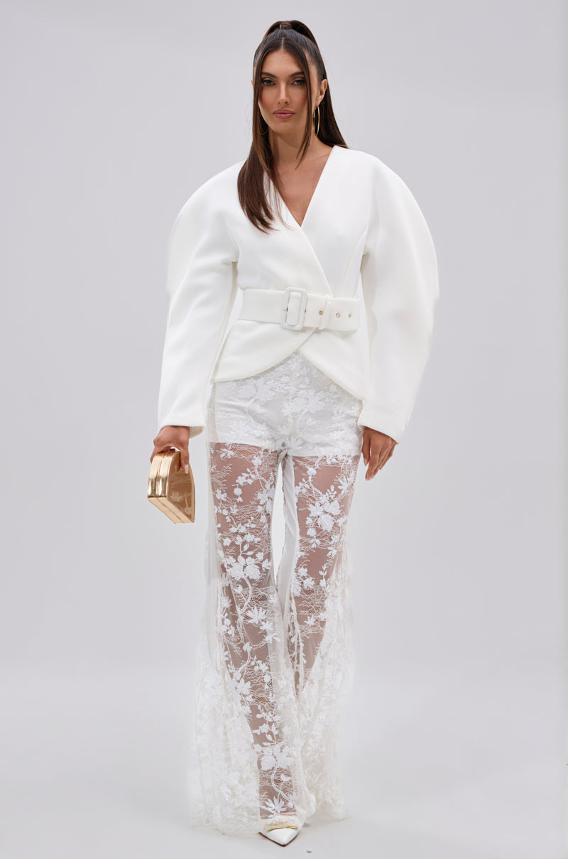 A woman poses in the LANA SCUBA BLOUSE with sculptural sleeves and a cinched waist, paired with sheer white lace pants, white pointed-toe heels, and a metallic gold clutch against a plain light gray background.