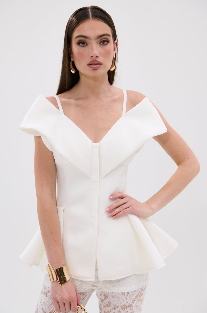 A woman in the DANCE AROUND SLEEVELESS PEPLUM CORSET—a chic white off-shoulder top with ruffles and peplum hem—paired with lace pants, stands against a plain background, accessorized with hoop earrings and a gold cuff bracelet. 1
