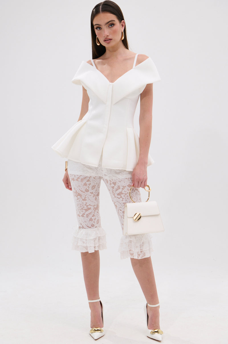 A woman poses against a white background wearing the DANCE AROUND SLEEVELESS PEPLUM CORSET, paired with white lace ruffled cropped pants, gold-tipped white heels, and holding a white handbag. 4