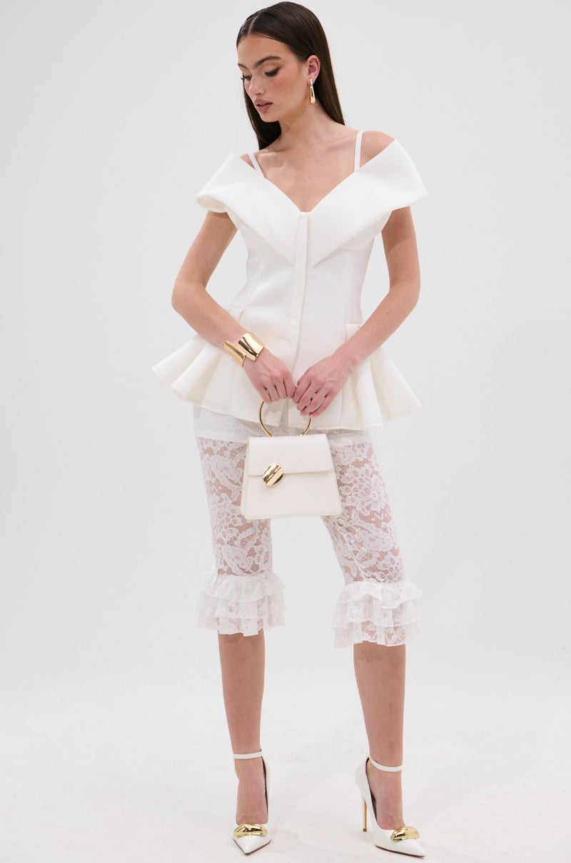 A woman wears the DANCE AROUND SLEEVELESS PEPLUM CORSET, paired with white lace ruffled capri pants, gold-tipped white heels, a small white handbag, gold hoop earrings, and has straight dark hair. 5
