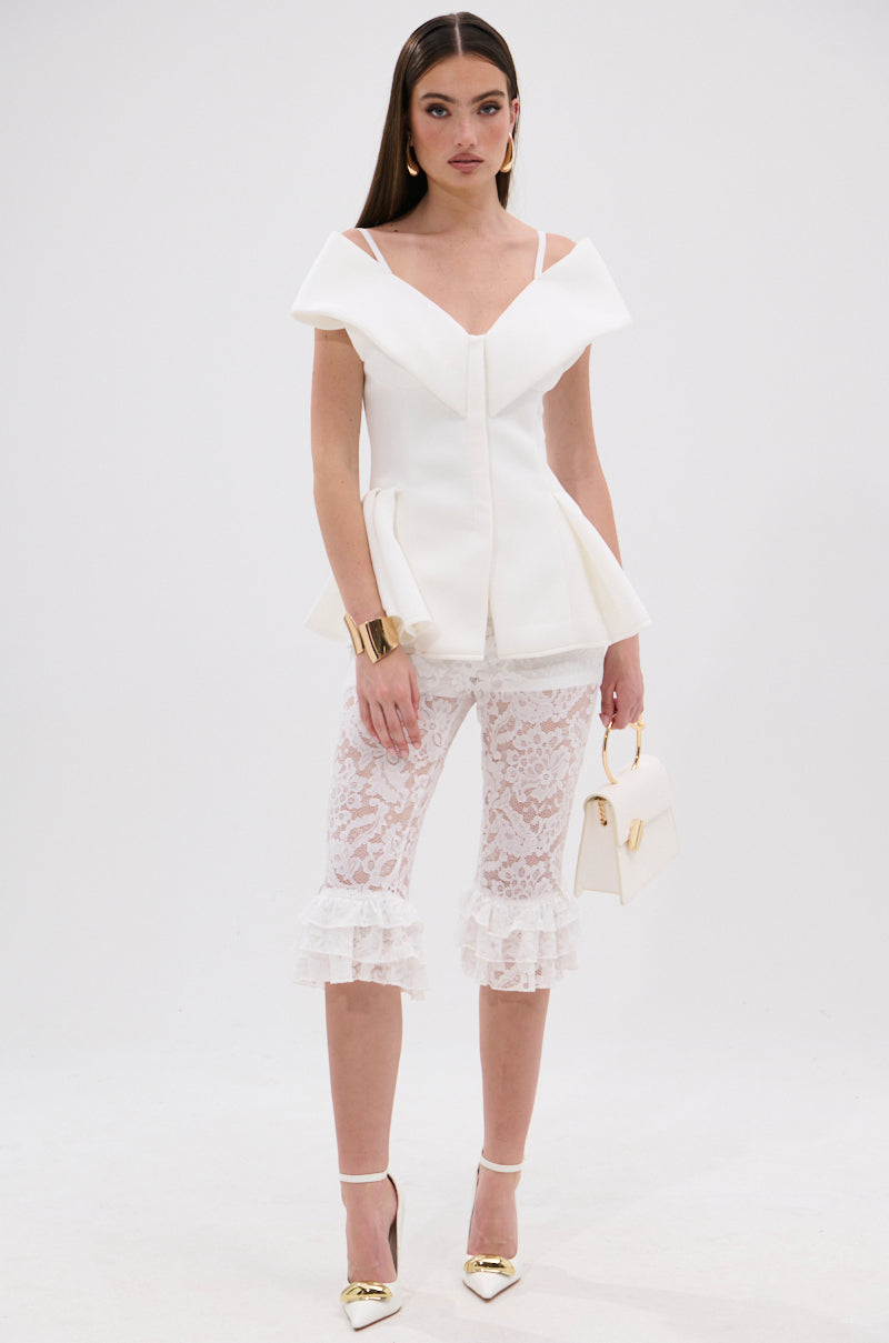 A woman stands against a white background wearing the DANCE AROUND SLEEVELESS PEPLUM CORSET, white lace ruffle pants, gold heels, a gold cuff bracelet, and holding a white handbag. 6