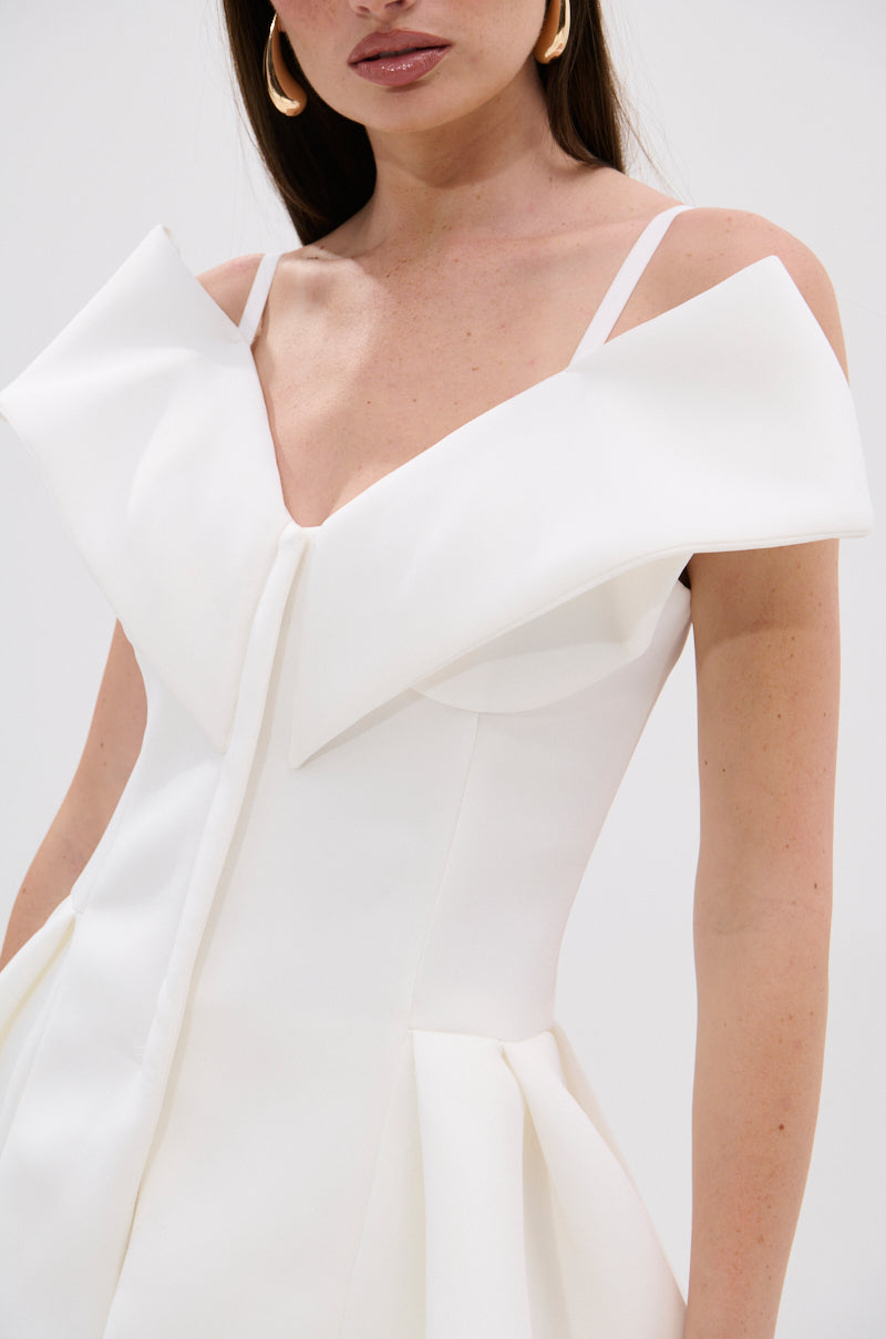 A woman wears the DANCE AROUND SLEEVELESS PEPLUM CORSET, a white dress with thin straps, a corset top, and a sculptural bow on the bodice. She accessorizes with gold hoop earrings against a plain, light background. 7