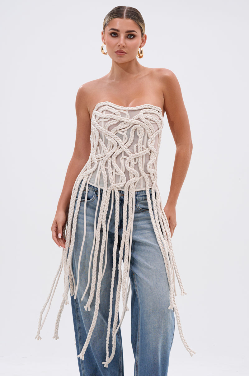 A woman with slicked-back hair and large hoop earrings wears the NEVER LOST TASSEL MESH CORSET—strapless with a sweetheart neckline, structural boning, and knotted rope details—paired with loose blue jeans against a plain background.