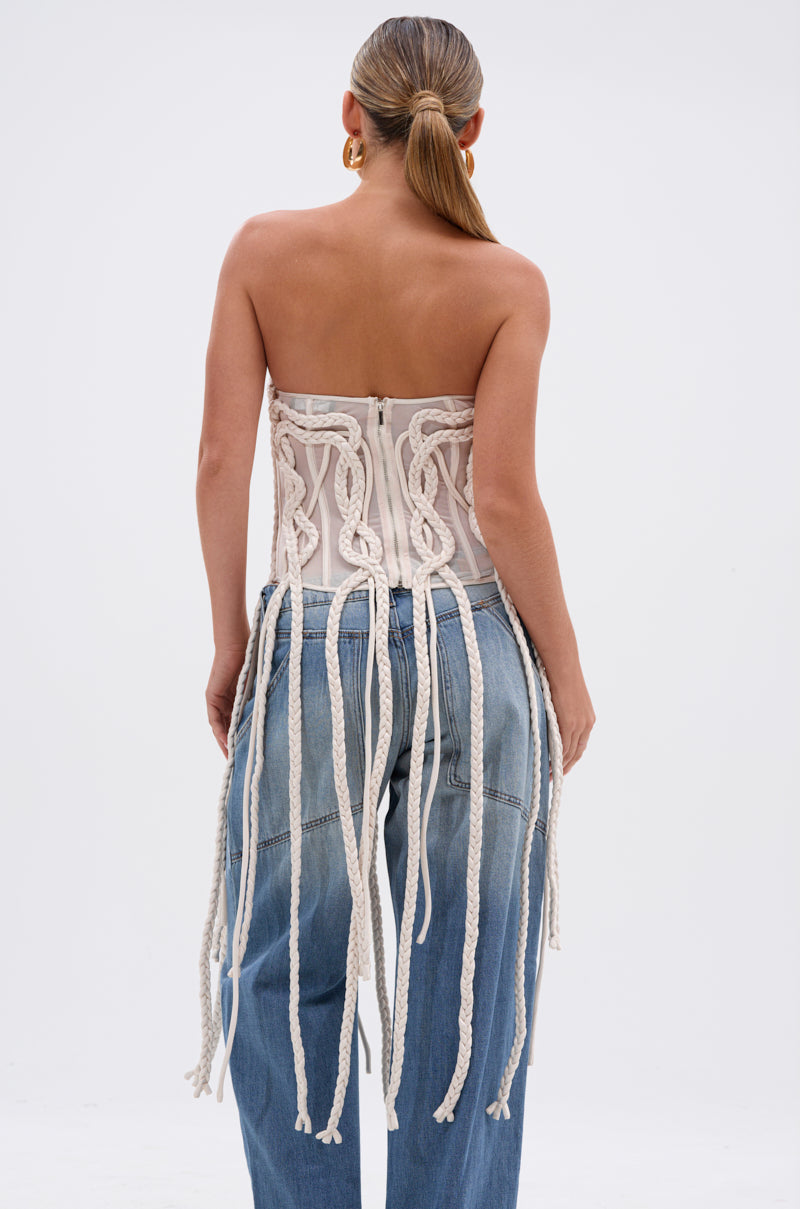 A woman with slicked-back hair and gold hoop earrings models the NEVER LOST TASSEL MESH CORSET, featuring a strapless sweetheart neckline, long braided tassels, and back details, paired with high-waisted blue jeans. She is shown from behind.