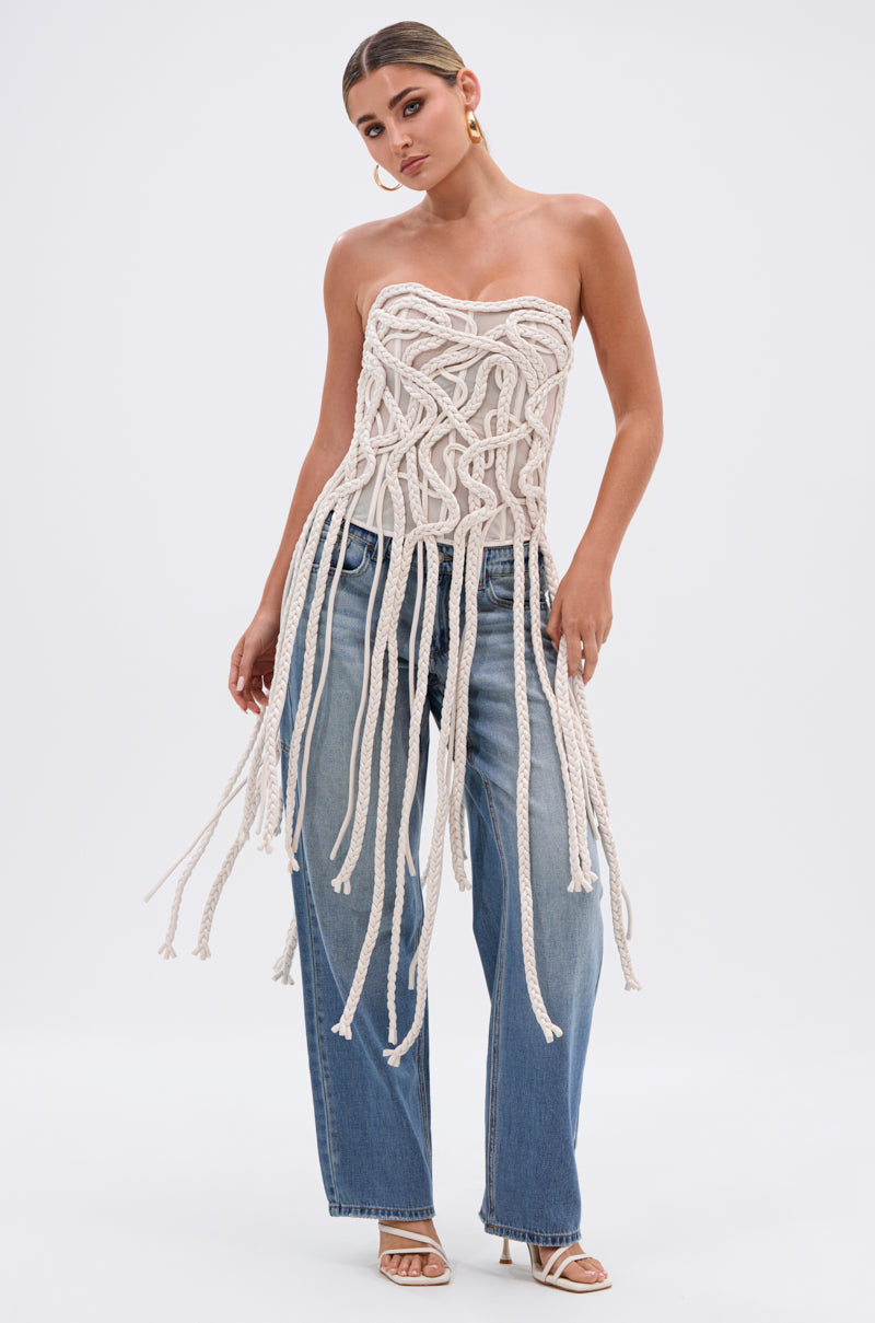 A woman poses in the NEVER LOST TASSEL MESH CORSET, a strapless cream top with a sweetheart neckline and long braided tassels, styled with wide-leg blue jeans, heeled sandals, hoop earrings, and slicked-back hair against a plain white background.