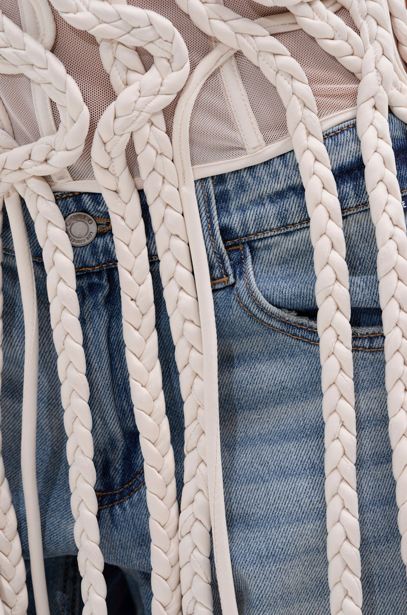 Close-up of a person in blue denim jeans wearing the NEVER LOST TASSEL MESH CORSET, detailed with long, thick white braided ropes draping elegantly over the jeans.