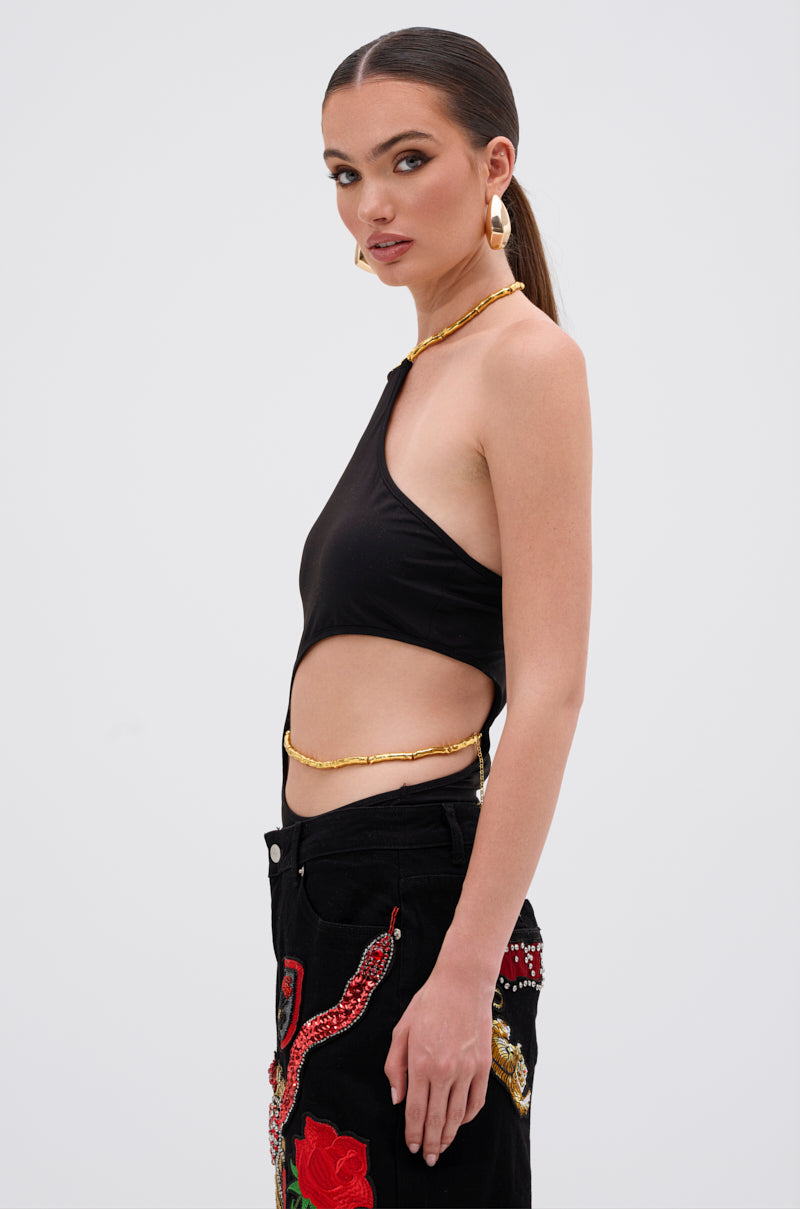 A woman with straight brown hair in a low ponytail wears the ALWAYS THINKING BODYSUIT WITH CUTOUT, gold jewelry, and black pants with red and gold designs, posing in profile against a plain background. 2