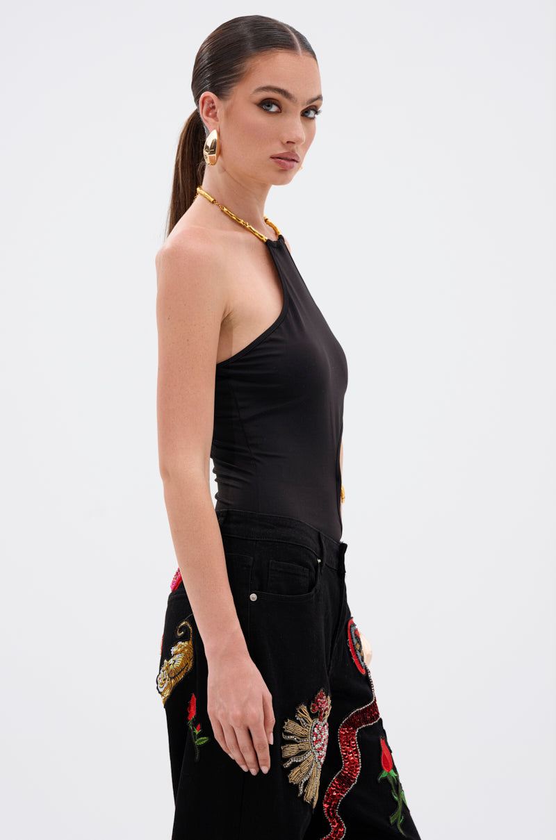 A woman with long brown hair in a sleek ponytail wears the ALWAYS THINKING BODYSUIT WITH CUTOUT—an elegant black one-shoulder style with sculptural side cutouts—paired with gold hoop earrings and colorful embroidered black pants against a white background. 3