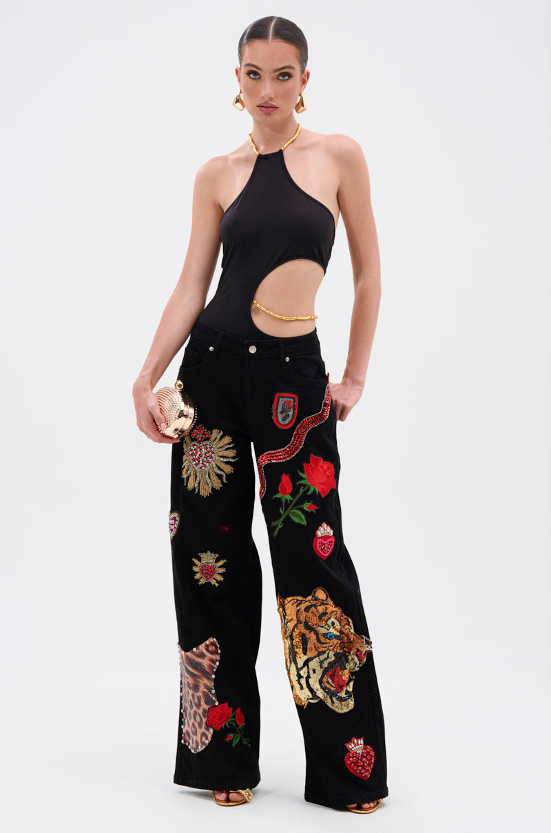A woman poses in the ALWAYS THINKING BODYSUIT WITH CUTOUT, a black halter-neck style with sculptural side cutouts, paired with wide-leg pants embroidered with tigers, roses, and hearts. She adds bold gold jewelry and a handbag with gold bamboo-style hardware. 5