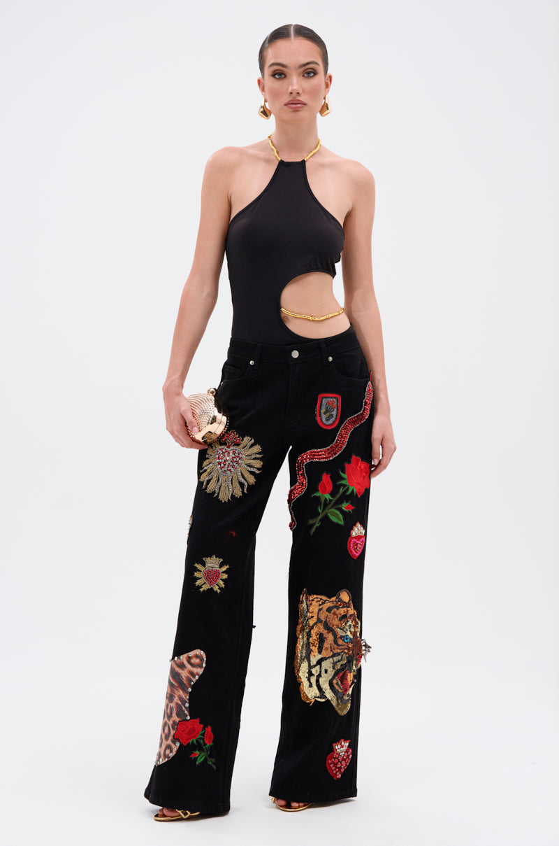 A woman poses against a plain background in the ALWAYS THINKING BODYSUIT WITH CUTOUT, a black halter-neck style with sculptural side cutouts, and high-waisted pants featuring embroidered animals, flowers, and hearts. She accessorizes with a small clutch and gold earrings. 6