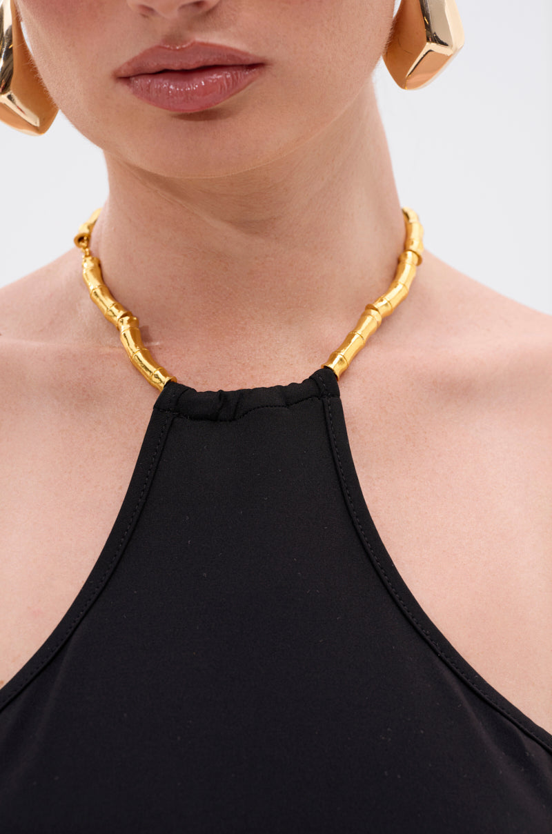 A close-up of a woman in the ALWAYS THINKING BODYSUIT WITH CUTOUT, featuring sleeveless black fabric with sculptural side cutouts, chunky gold hoop earrings, and a gold bamboo-style necklace. The image is cropped just below her nose. 7