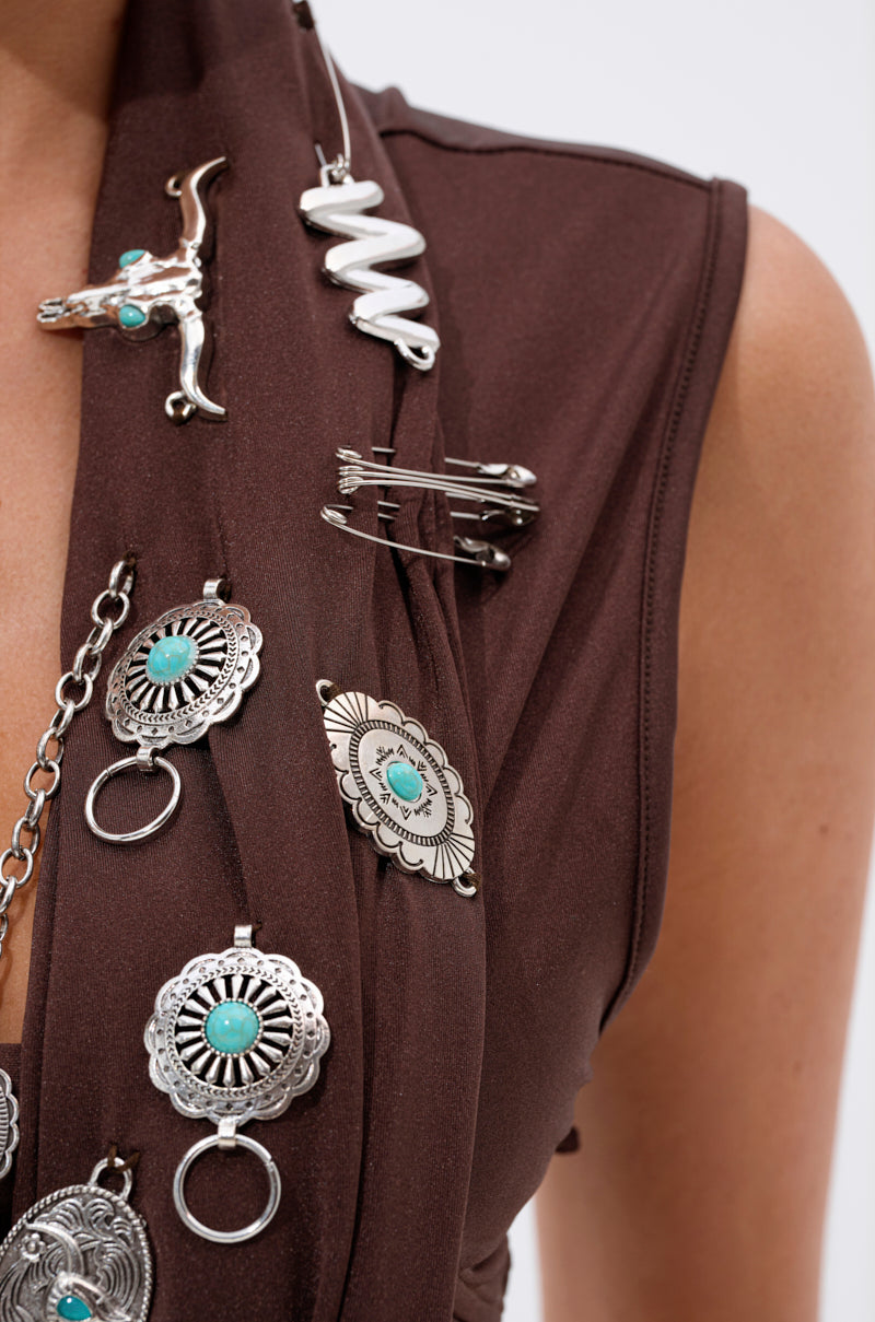 A close-up of a person wearing the SO BLESSED COWL NECK BLOUSE, a cropped sleeveless top decorated with silver pins, brooches, turquoise stones, and Western-themed circular, floral, and gun-shaped charms. 11