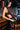 A woman with long dark hair leans against a wooden bar, wearing the SO BLESSED COWL NECK BLOUSE and a black and brown patterned skirt. The background features a rustic bar interior.