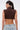 A woman with long dark hair in a ponytail wears large hoop earrings, light blue jeans, and the SO BLESSED COWL NECK BLOUSE in brown. She stands with her back to the camera against a plain background, radiating Western-inspired charm.