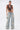 A woman poses against a plain background, wearing the SO BLESSED COWL NECK BLOUSE styled with light blue wide-leg distressed jeans, silver accessories, white heels, and a small silver purse featuring Western-inspired charms.