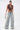 A woman poses against a white background, wearing the SO BLESSED COWL NECK BLOUSE and light blue ripped wide-leg jeans. She holds a small silver beaded purse and has her hair slicked back.
