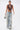 A woman in the SO BLESSED COWL NECK BLOUSE, wide-leg distressed jeans, bold necklaces, hoop earrings, and a small silver handbag walks forward against a white background.