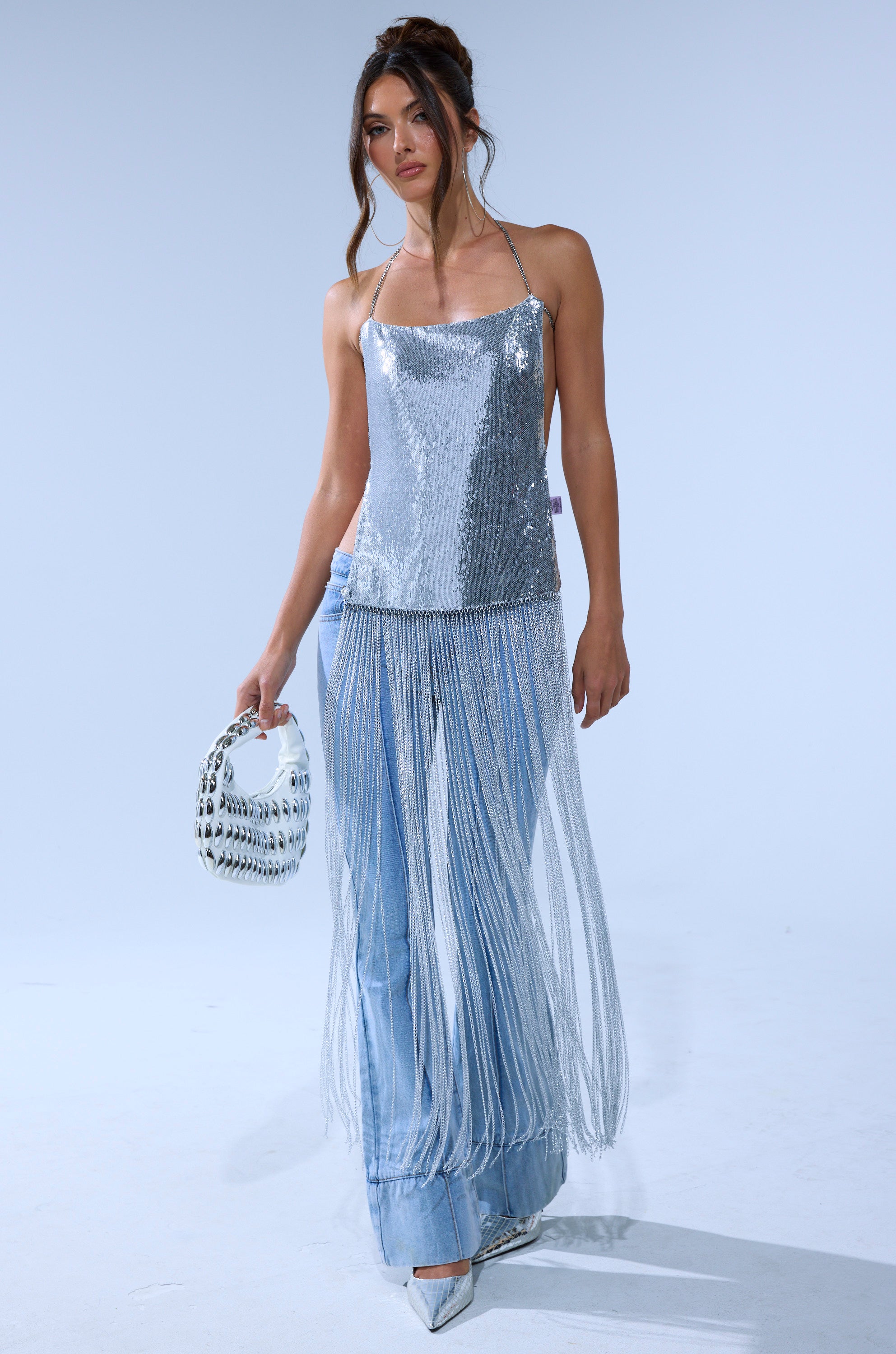 A woman models the ONLY THINK ABOUT YOU SILVER RHINESTONE FRINGE TOP with a halter neckline, pairing it with light blue jeans, silver pointed-toe heels, and a round silver handbag against a plain light background.