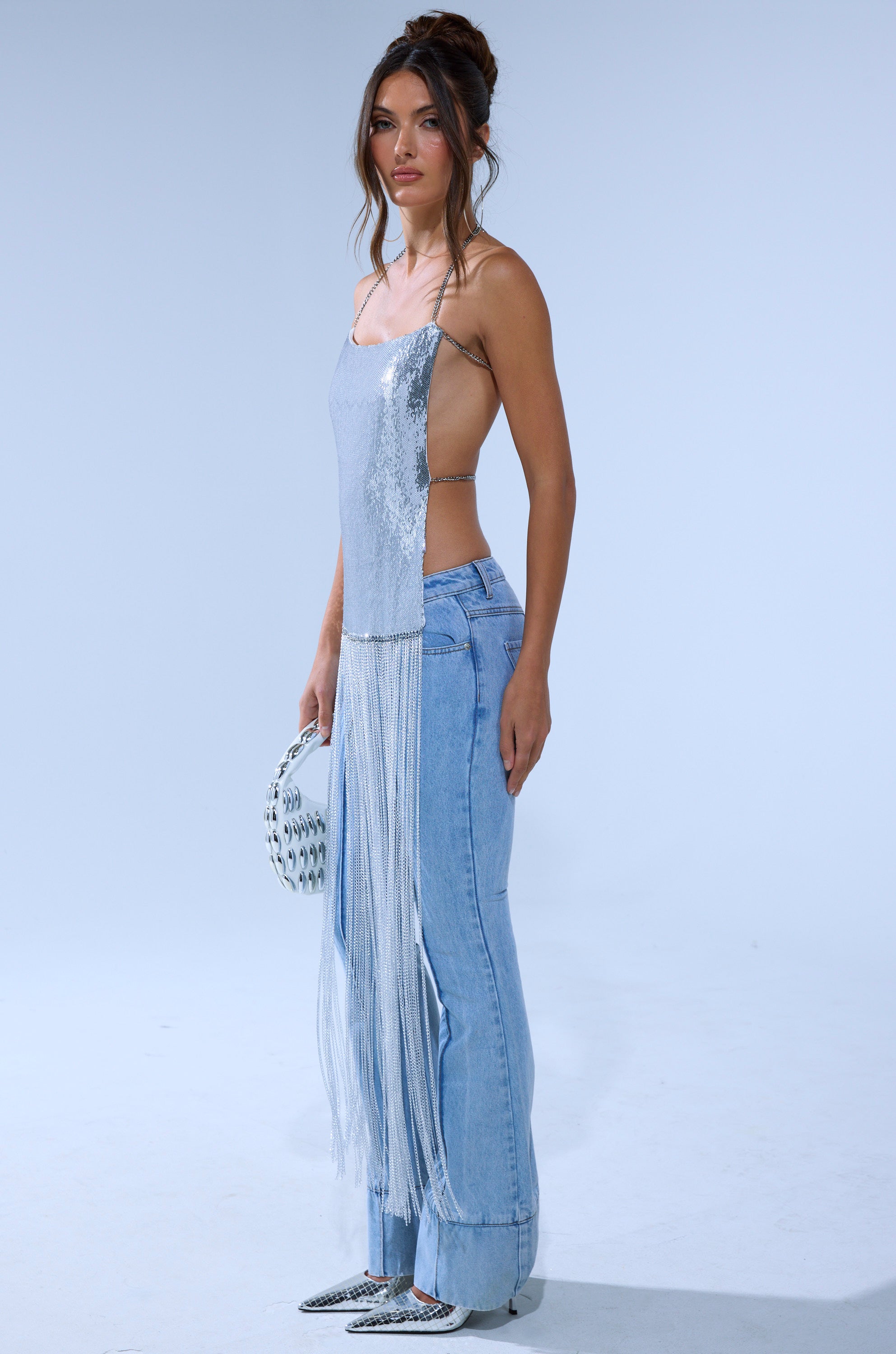 A woman in profile wears the ONLY THINK ABOUT YOU SILVER RHINESTONE FRINGE TOP, styled with light blue high-waisted jeans, silver pointed shoes, and a white clutch with metal details against a plain light background.