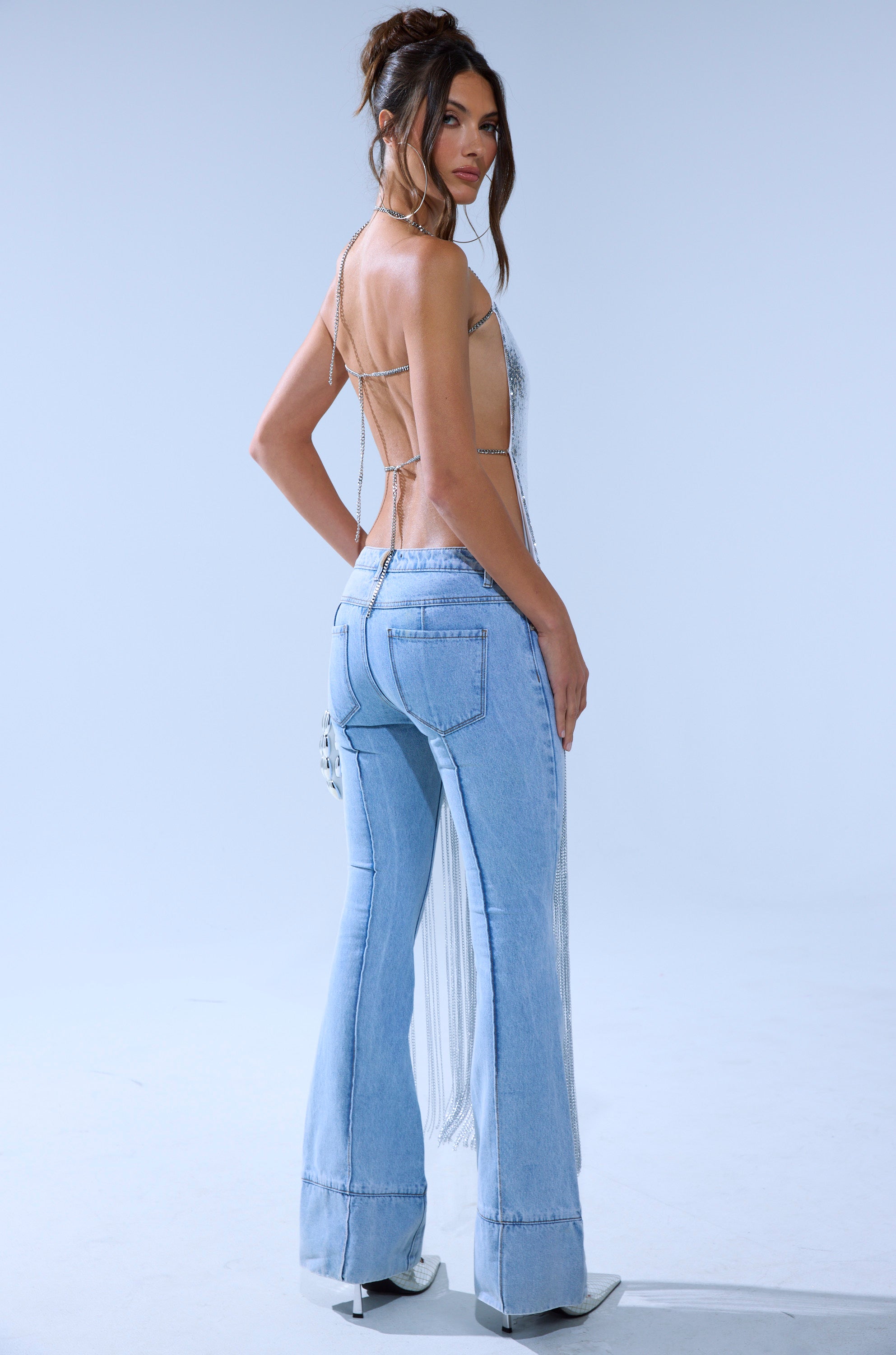 A woman stands turned slightly over her shoulder, wearing light blue flared jeans, white pointed-toe shoes, and the ONLY THINK ABOUT YOU SILVER RHINESTONE FRINGE TOP with a halter neckline, set against a plain light background.