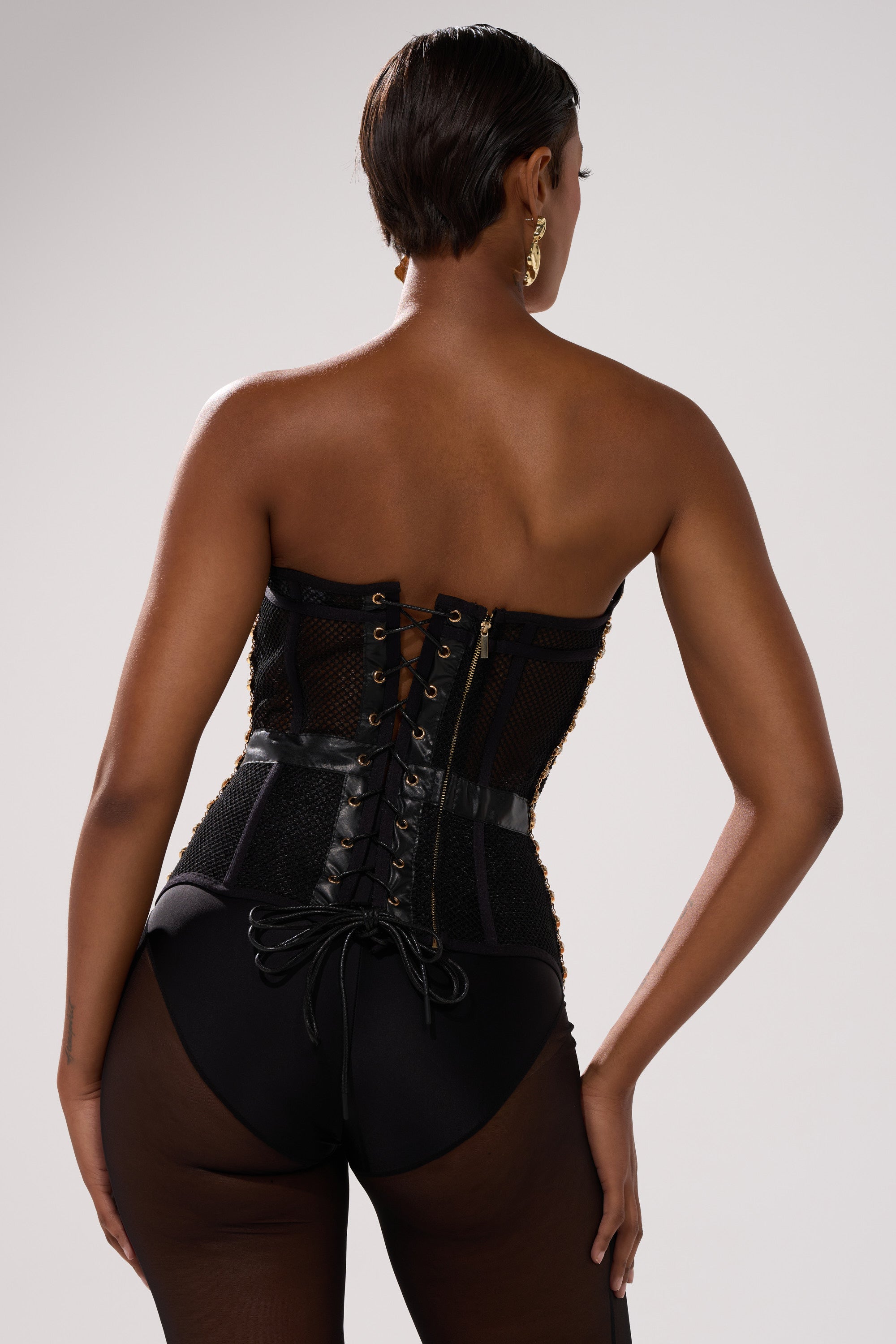 A person with short hair is shown from the back wearing a black Smokeshow Corset with rhinestones and lace-up details, paired with black sheer tights, against a neutral background.