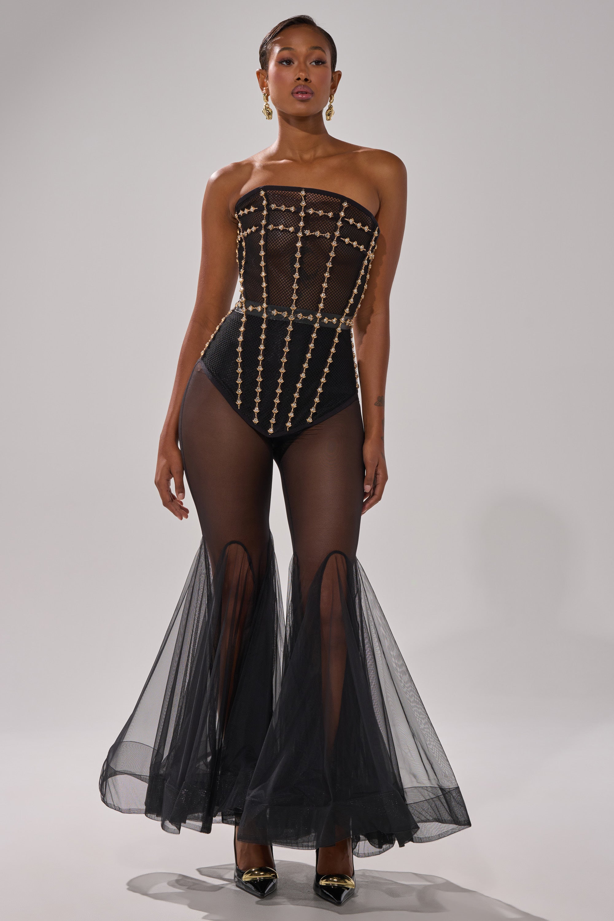 A woman wears the SMOKESHOW CORSET WITH RHINESTONES, a strapless black bodysuit with gold embellishments, paired with sheer black flared tulle pants, black pointed heels, and gold earrings against a plain light background.
