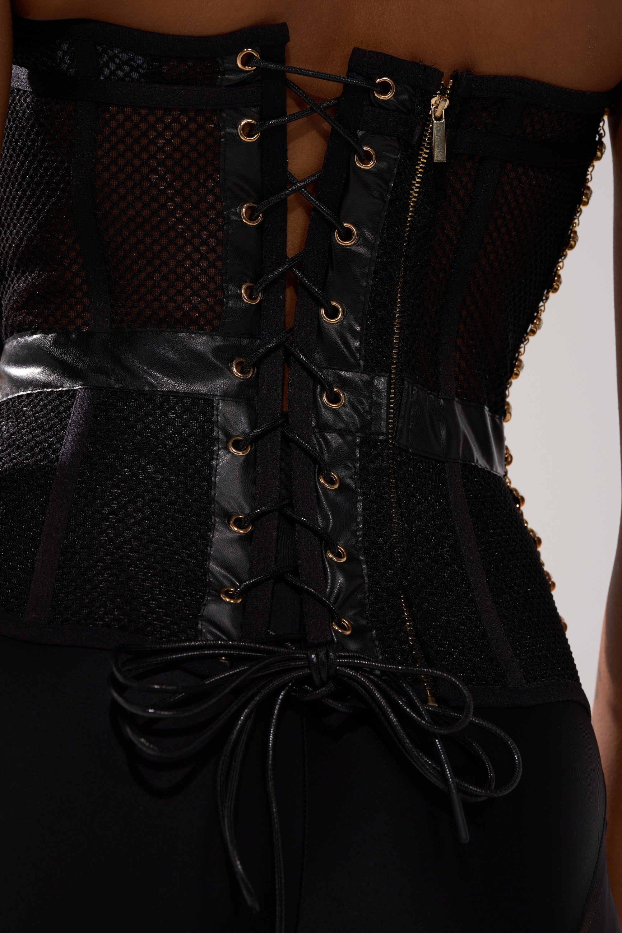 Close-up of a person wearing the SMOKESHOW Corset with Rhinestones, featuring sheer mesh, leather accents, gold eyelets, a lace-up back with laces tied in a bow, and a side zipper.