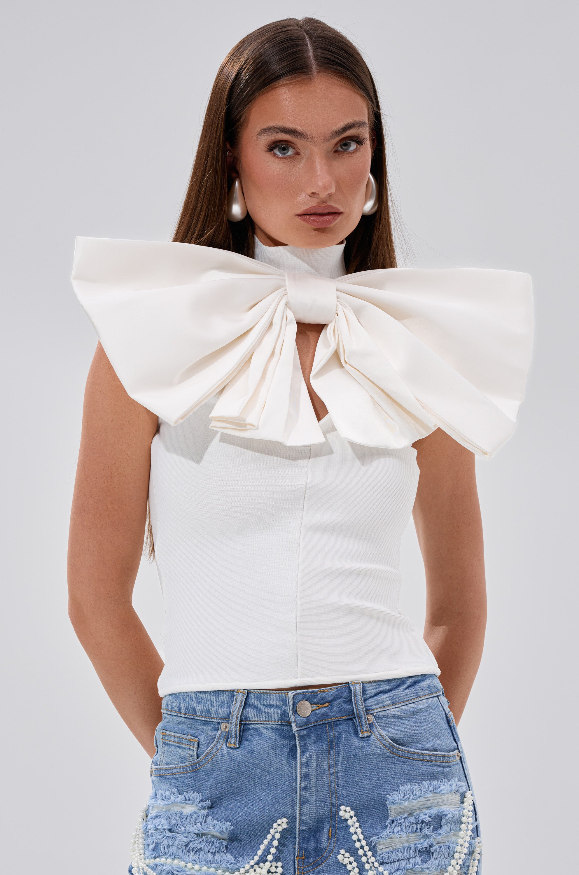 A woman styles her long straight brown hair and large white hoop earrings with the CUTIE BOW TOP IN WHITE—a sleeveless bandage top featuring a bold neck bow—plus pearl-embellished blue jeans, creating a chic ensemble.