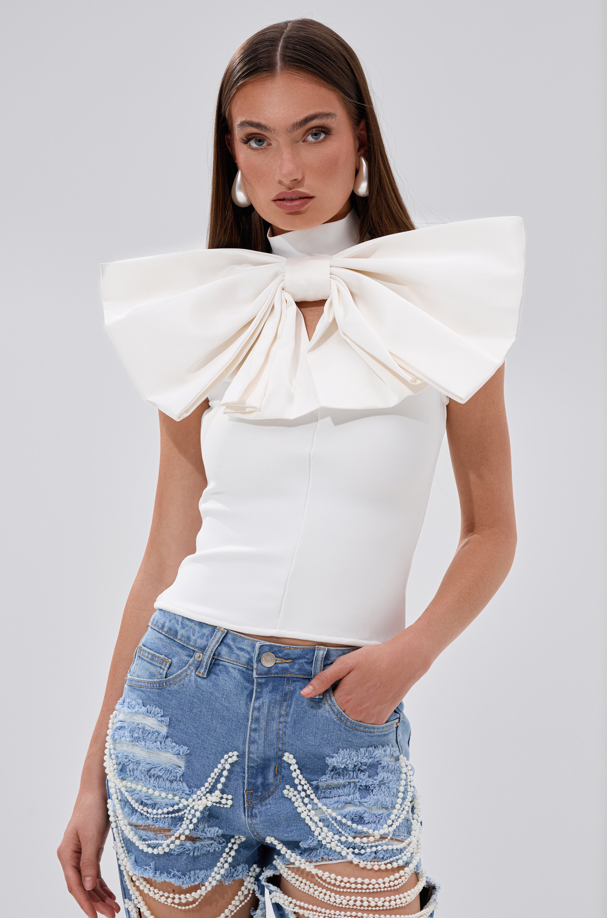 A woman confidently poses against a plain light background wearing the CUTIE BOW TOP IN WHITE—an oversized bow top—paired with ripped denim shorts featuring draped pearl chains.