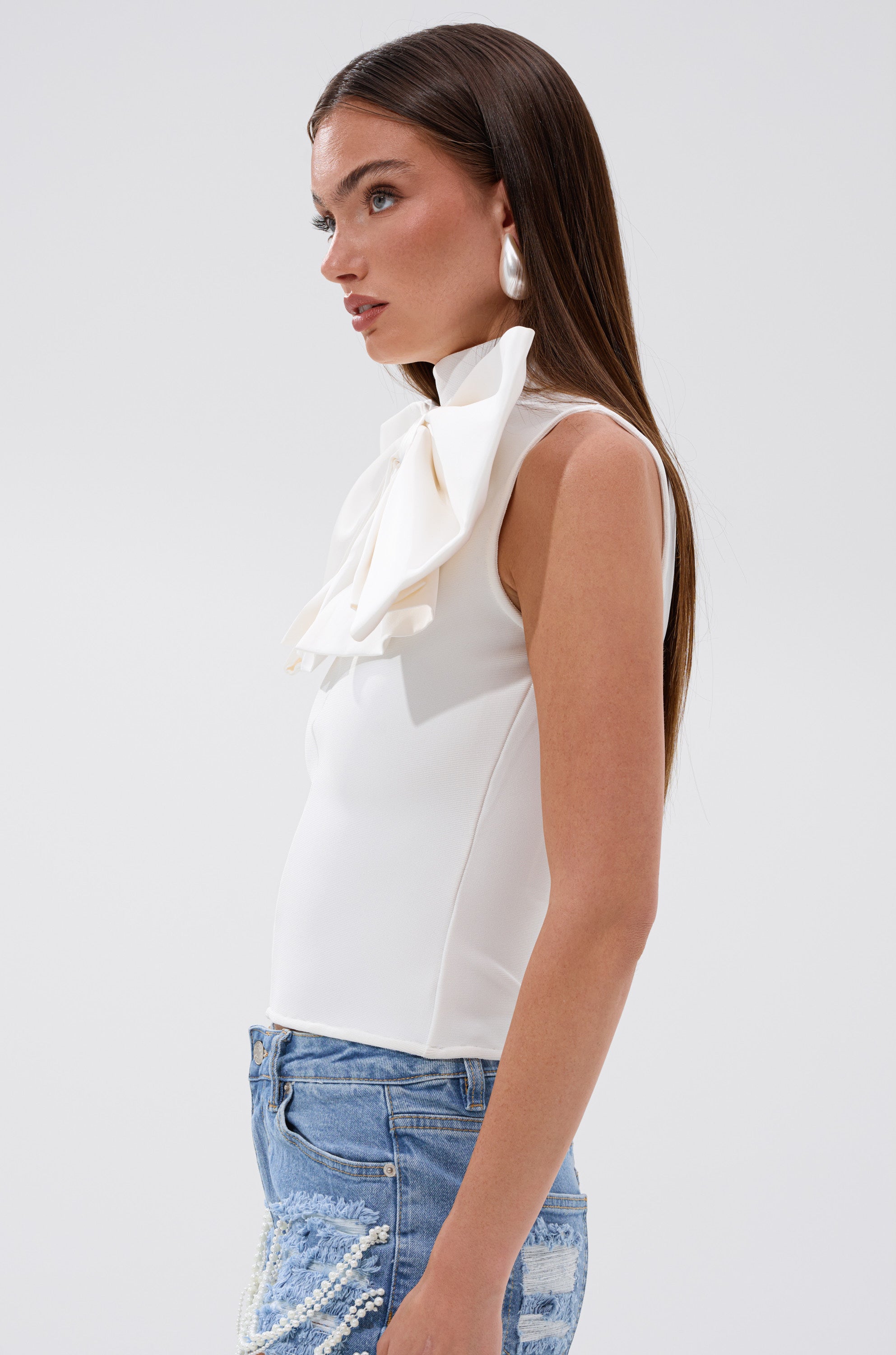Woman in the CUTIE BOW TOP IN WHITE, a sleeveless top with an oversized neck bow, paired with pearl earrings and blue beaded-detail jeans, stands in profile against a plain light background.