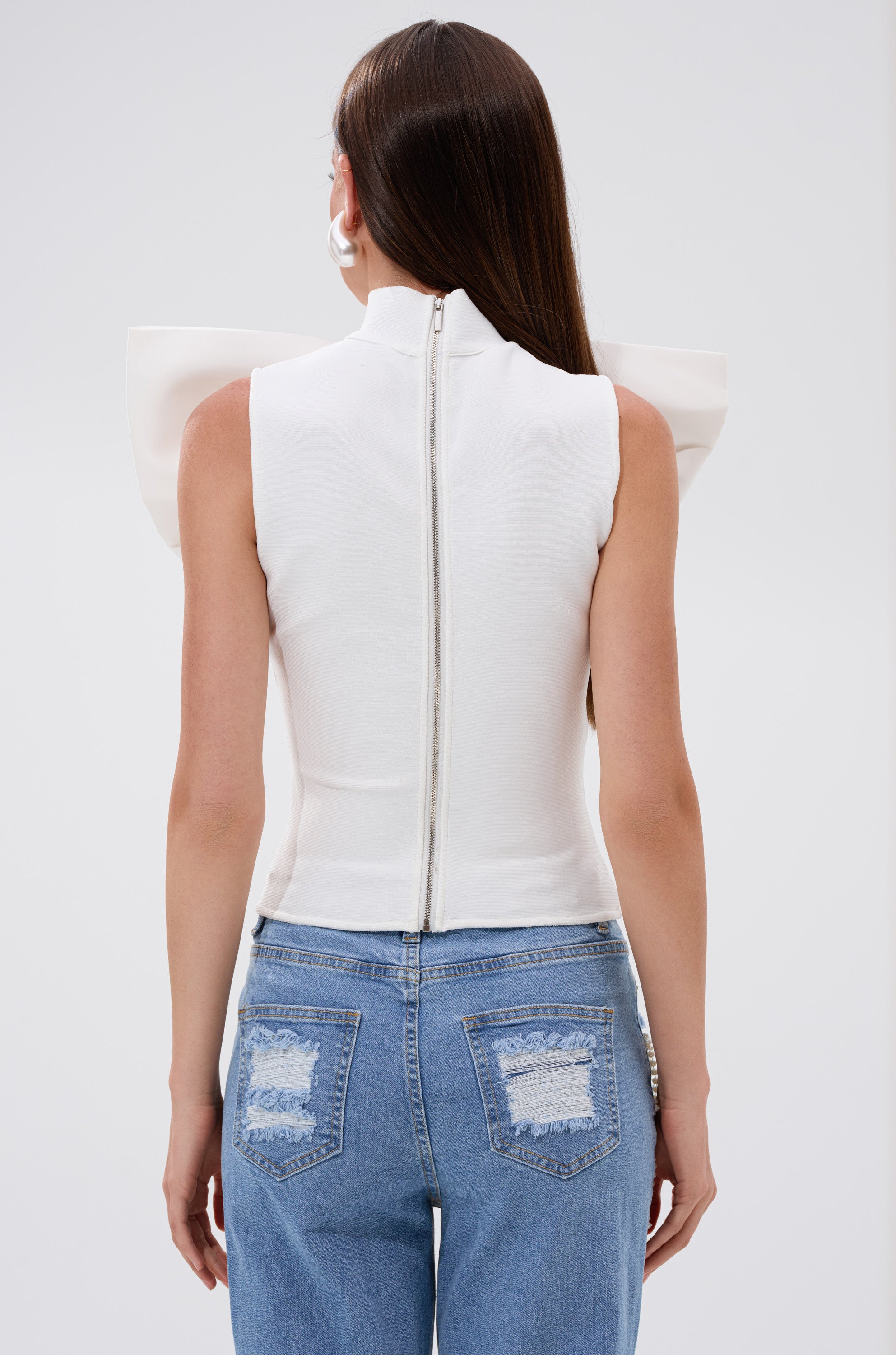A woman with long brown hair, facing away, wears the CUTIE BOW TOP IN WHITE with structured shoulders and a visible back zipper, paired with blue jeans featuring distressed back pocket patches.