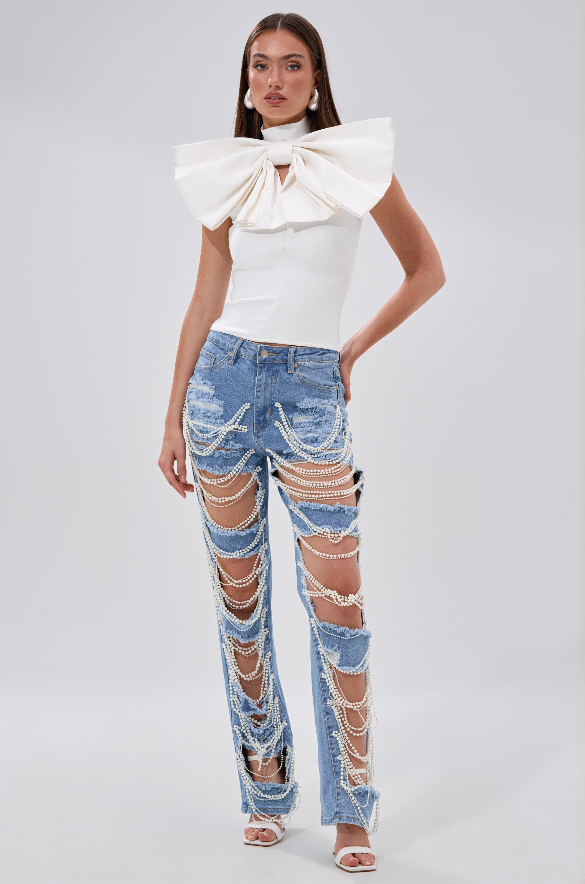 A woman models the CUTIE BOW TOP IN WHITE, a sleeveless body-contouring top, paired with distressed blue jeans adorned with pearl chains and white heeled sandals, standing against a plain background.