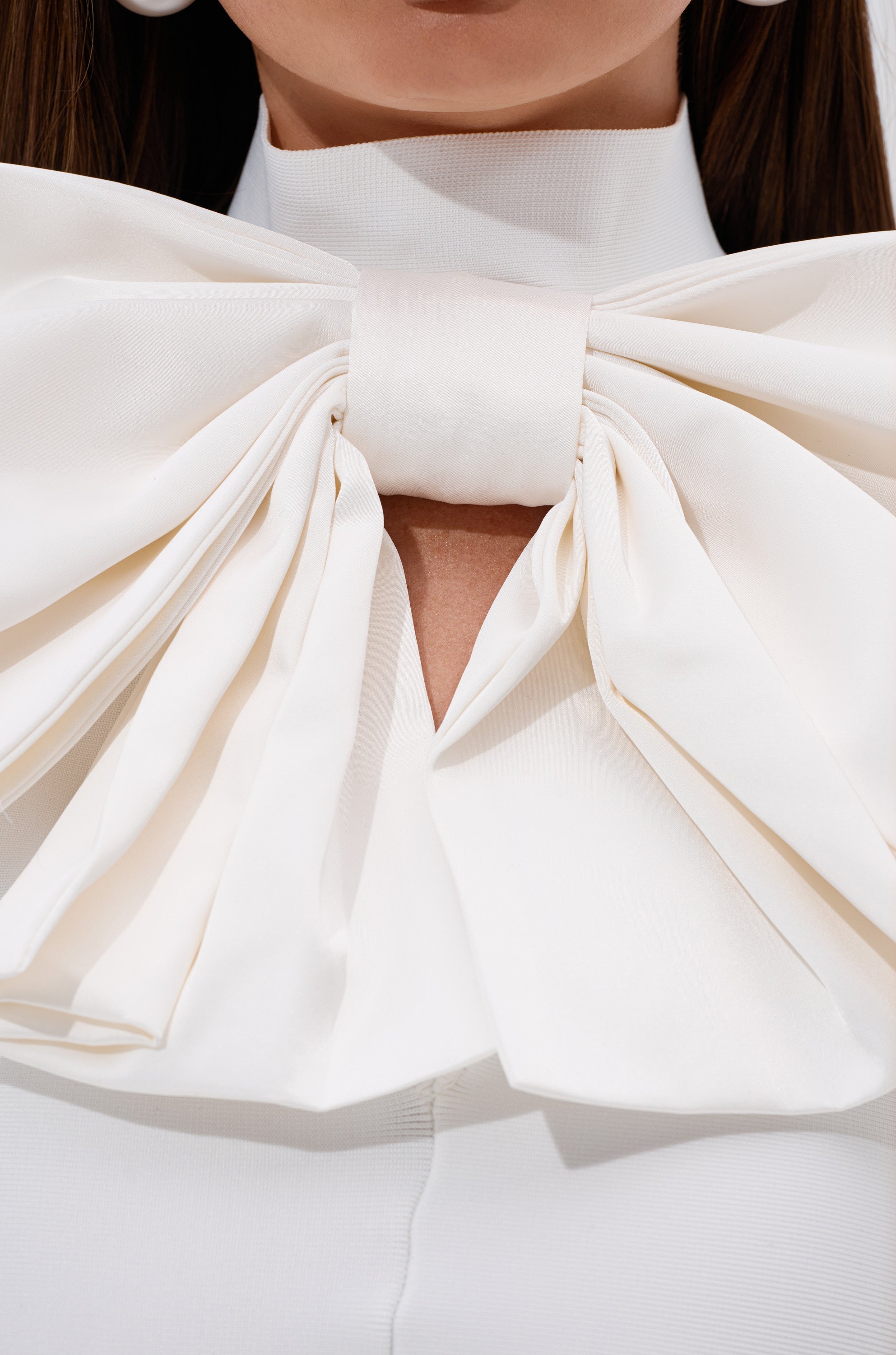 A close-up of a woman wearing the CUTIE BOW TOP IN WHITE, featuring a large, elegant satin bow tied at her neck over a high-collared white garment. The voluminous bow adds sophistication to her look.