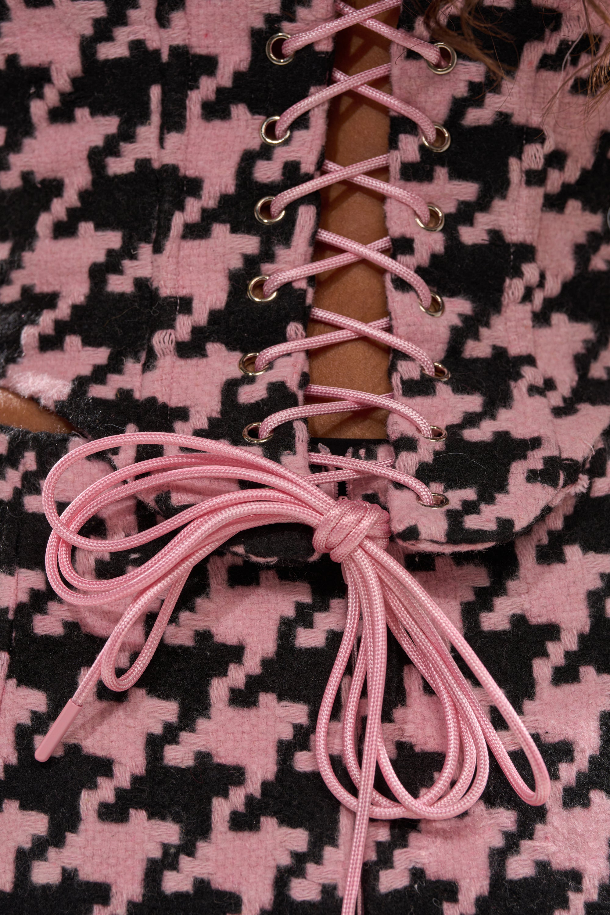 Close-up of the SMOOTH TRANSITION CORSET, showcasing pink and black houndstooth fabric, a lace-up front with metal eyelets, pink ribbon tied in a bow, and delicate satin flower details. 9