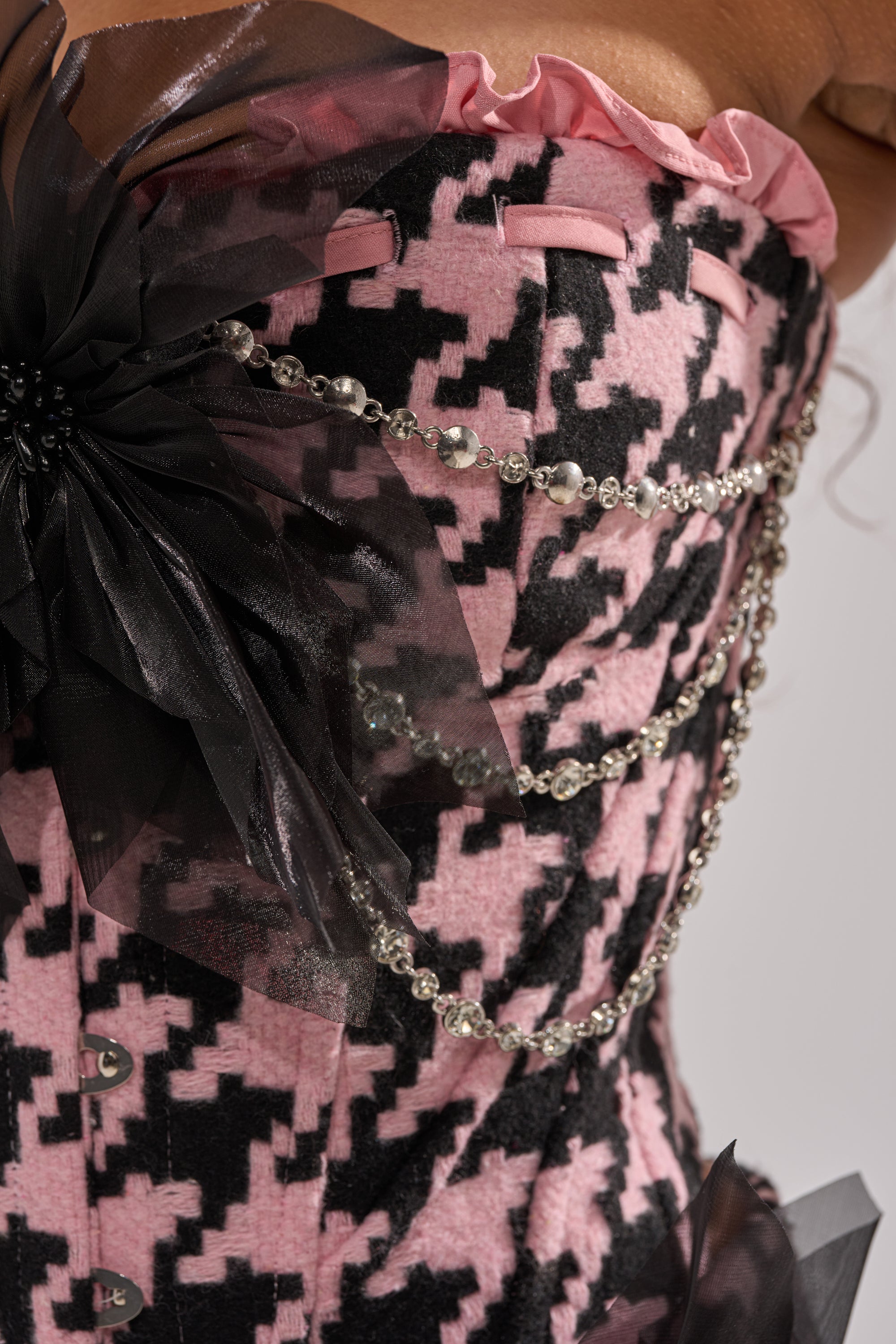 A close-up of the SMOOTH TRANSITION CORSET featuring a pink and black houndstooth pattern, sheer black fabric, satin flower accents, and draped silver beaded chains. 7