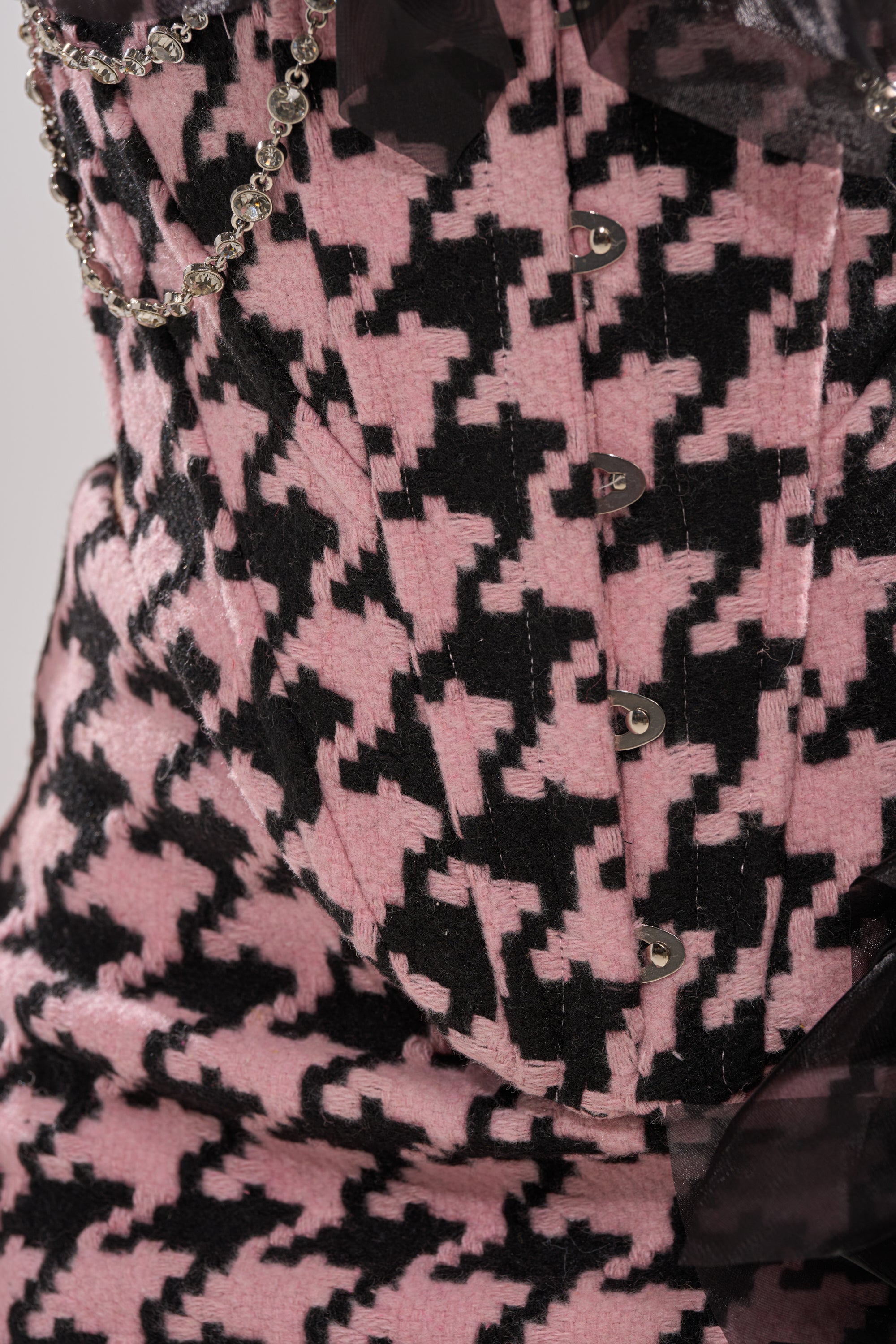 Close-up of the SMOOTH TRANSITION CORSET featuring a pink and black houndstooth pattern, decorative metal clasps and chain, corset-inspired structure, black sheer fabric accents, and elegant satin flower details. 8