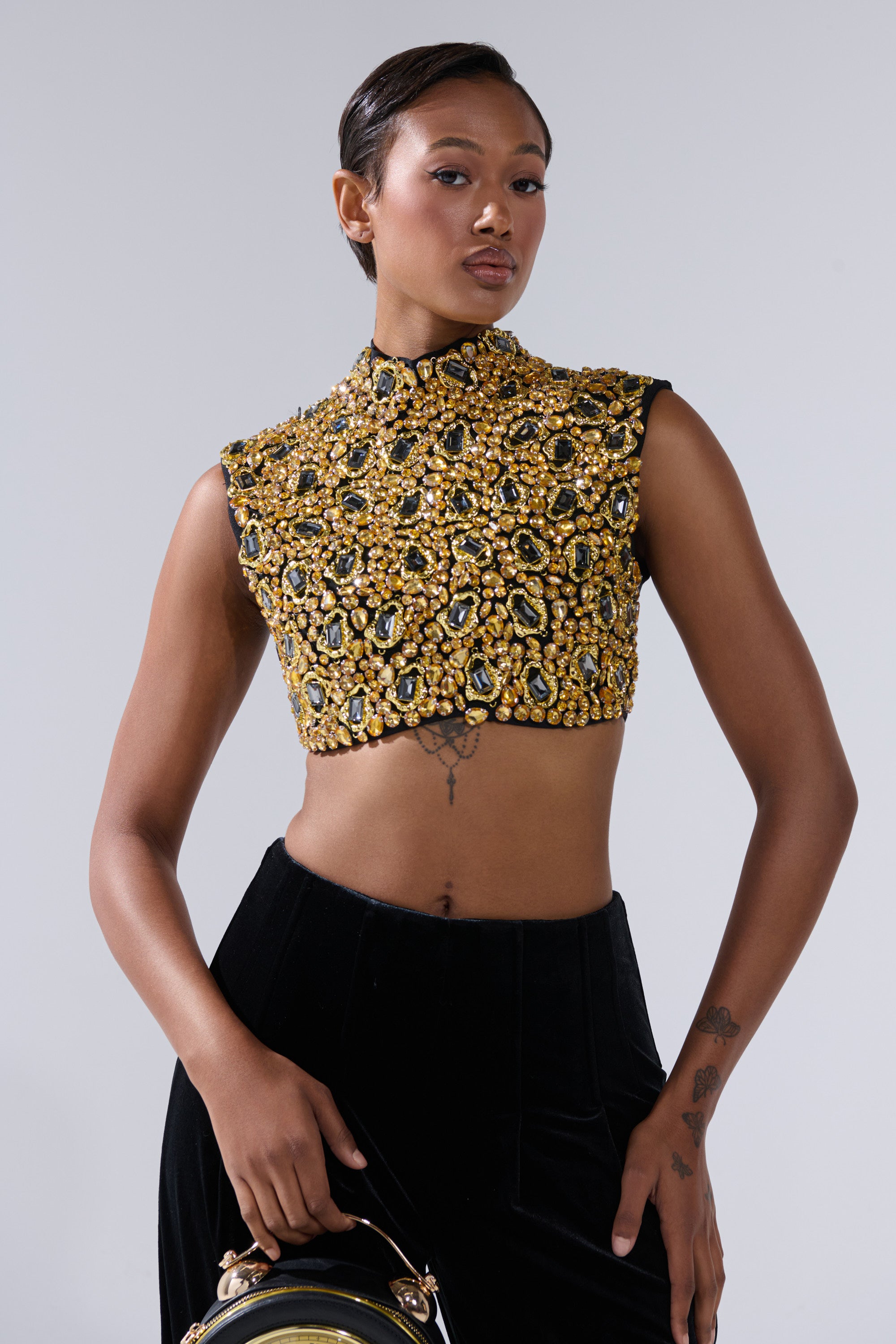 A person stands confidently wearing the GOLDEN GLAM BLOUSE with high black pants, holding a round black bag. Their slicked-back hair and left forearm tattoos complete the look.