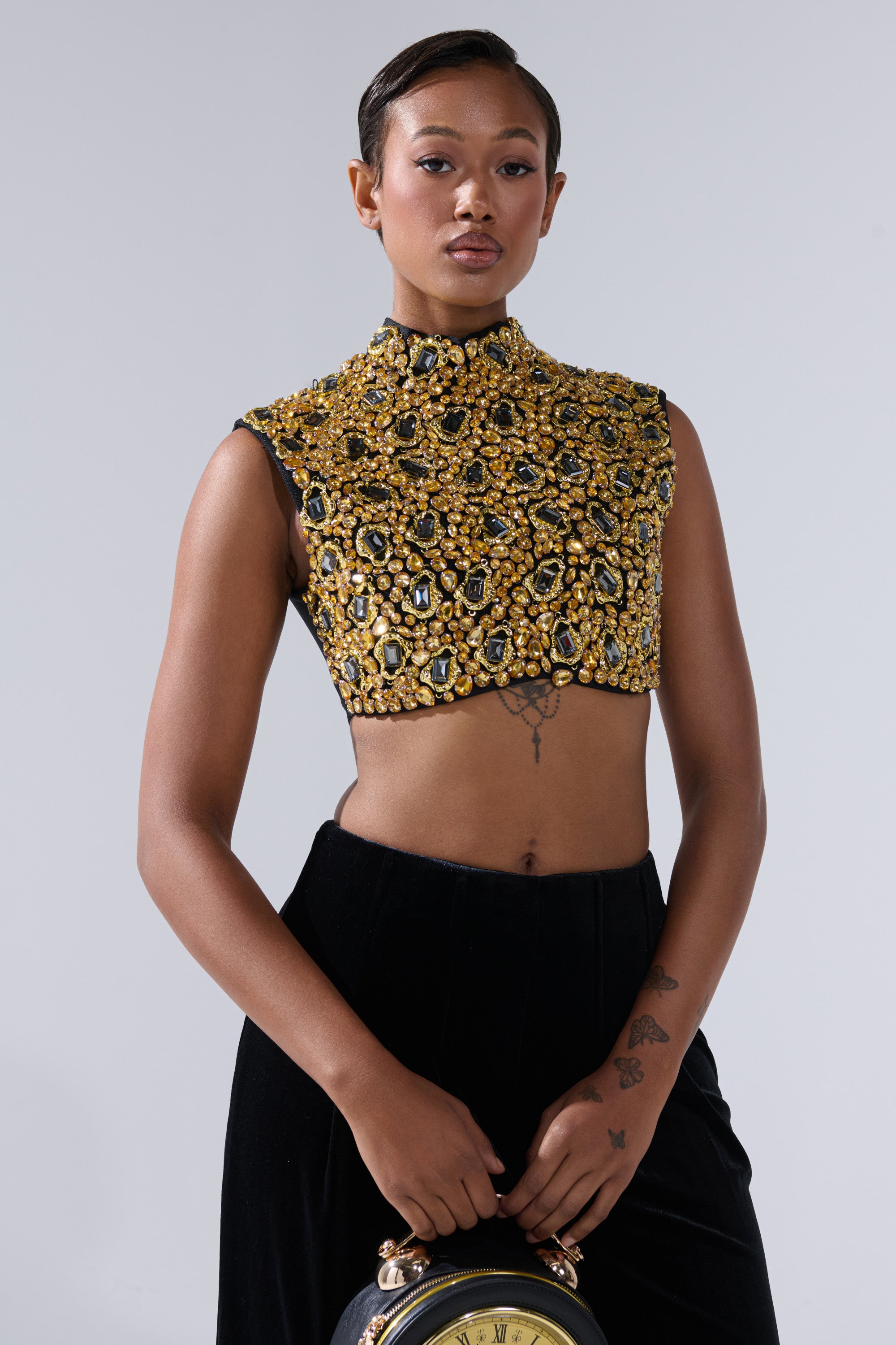 Wearing the GOLDEN GLAM BLOUSE with a high neck and sleeveless design, a short-haired individual with visible arm and torso tattoos pairs it with black pants and holds a round clock-inspired bag.