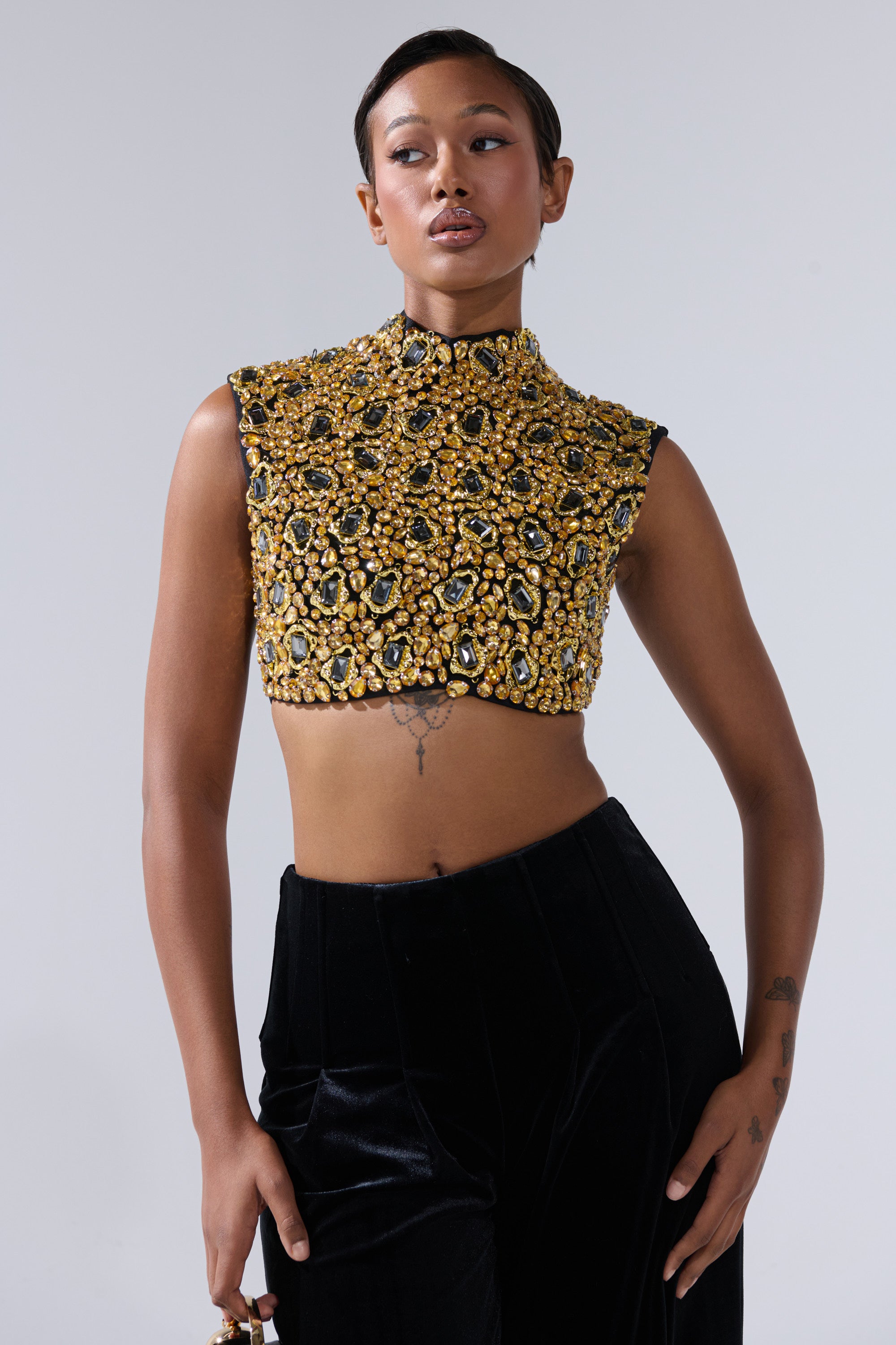 Wearing the GOLDEN GLAM BLOUSE—sleeveless, high-neck, with jeweled accents—paired with high-waisted black velvet pants, a person stands against a plain light background, gazing slightly to the side.