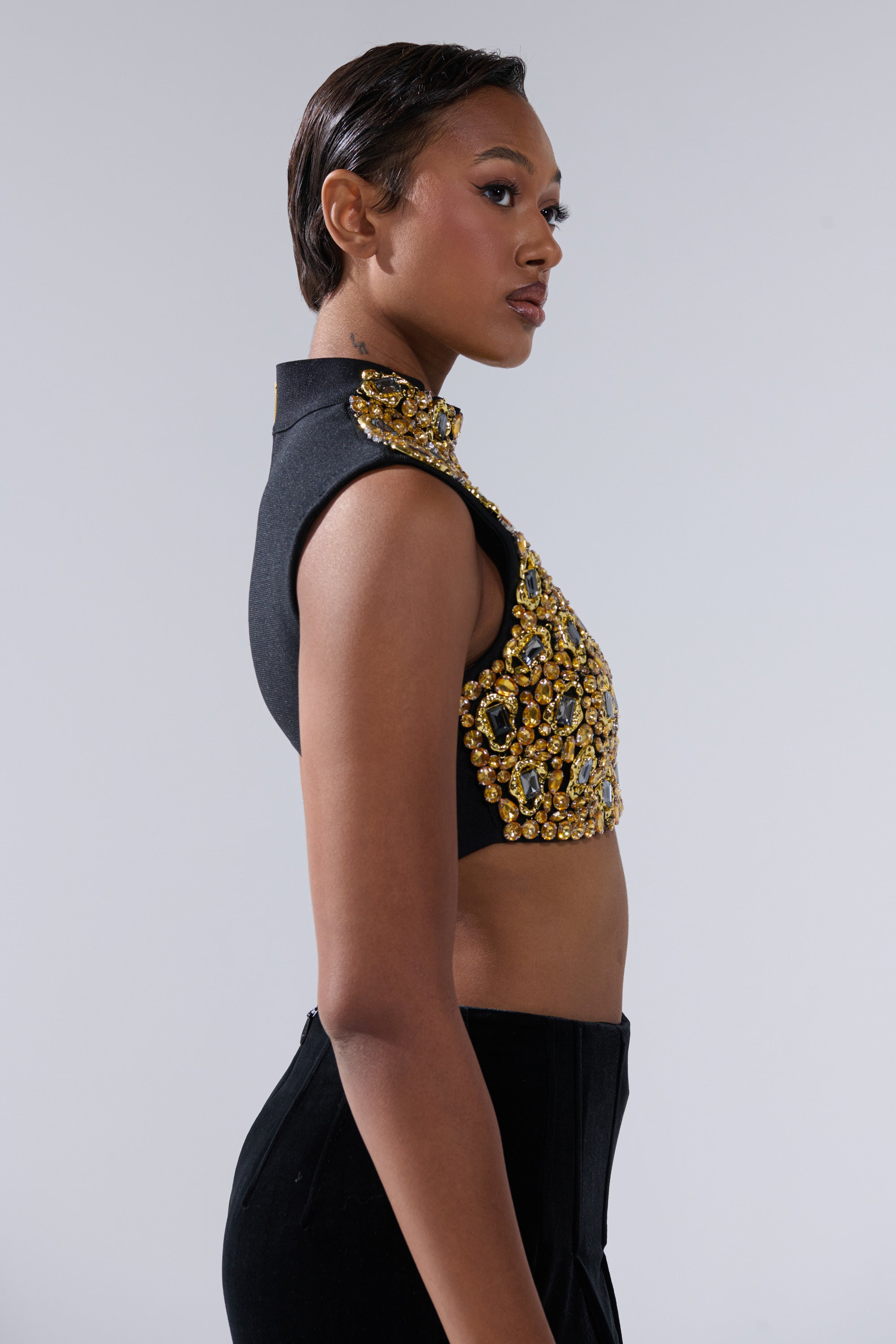 A woman in profile against a plain background wears the GOLDEN GLAM BLOUSE—a sleeveless black crop top with gold and jeweled embellishments—paired with high-waisted black pants and her hair slicked back.