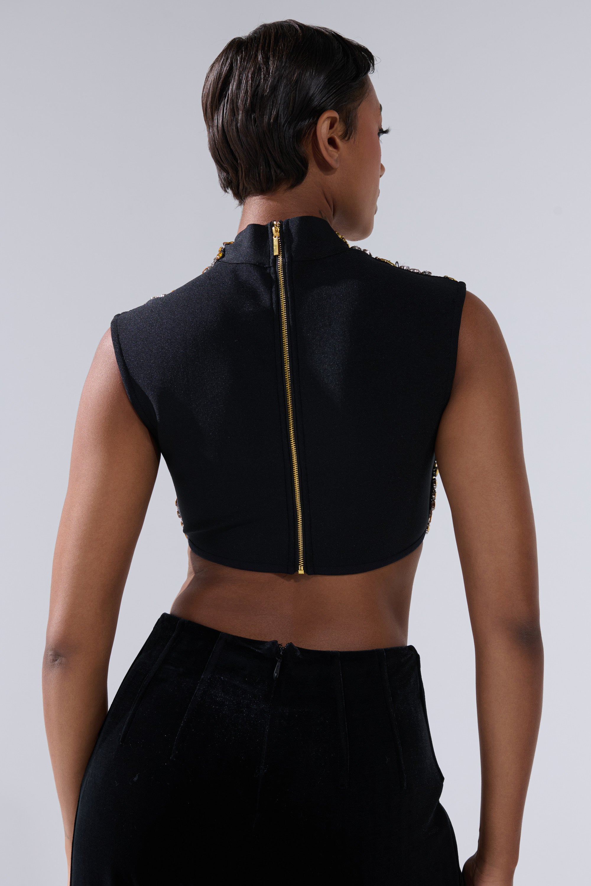 A person with short dark hair is seen from behind, wearing the GOLDEN GLAM BLOUSE—a sleeveless black top with a gold zipper—paired with high-waisted black pants against a plain light background.
