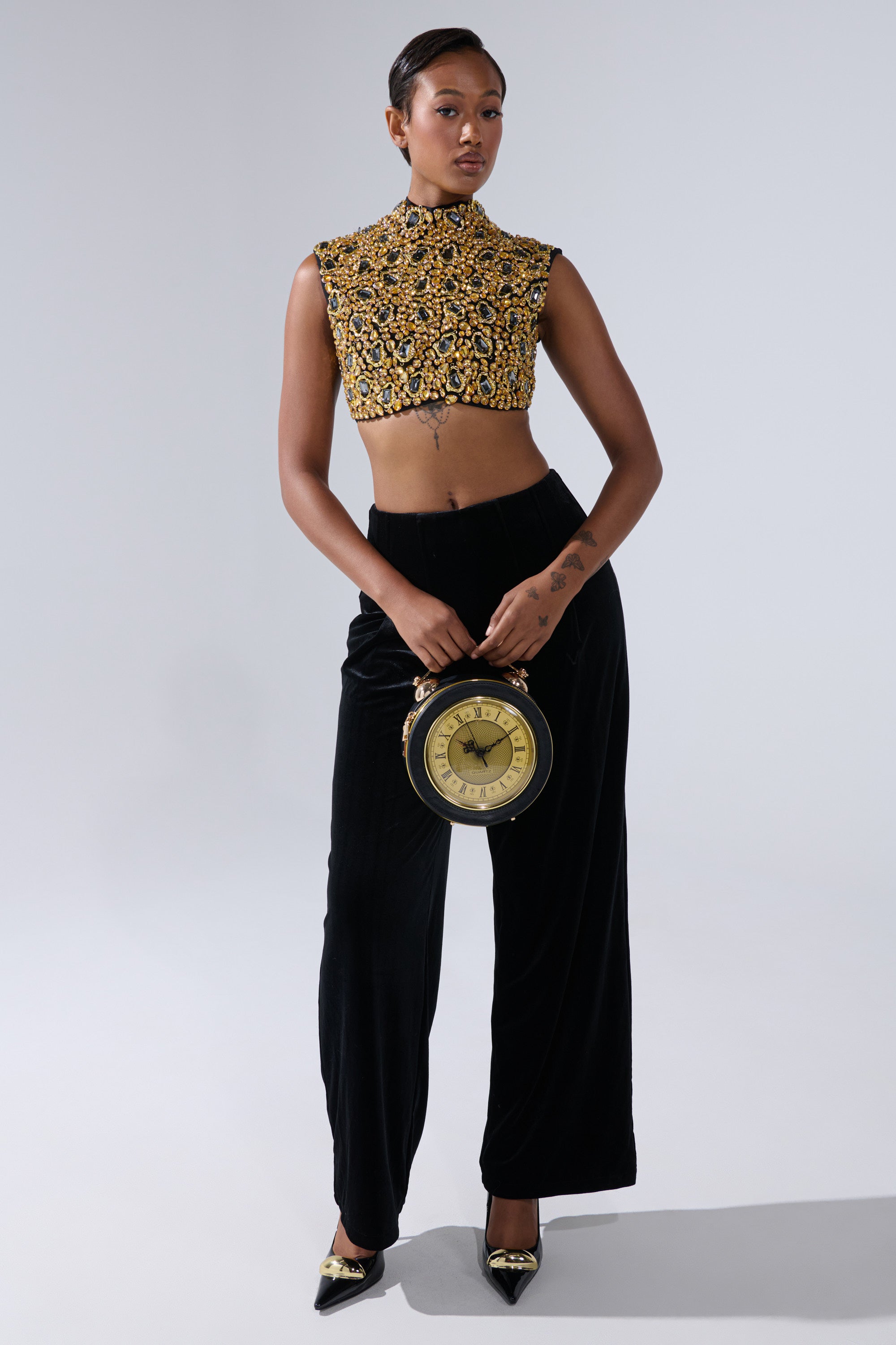 A woman stands confidently in the GOLDEN GLAM BLOUSE and black velvet pants, holding a large clock by her side. She wears black pointed heels with gold embellishments against a plain light gray background.