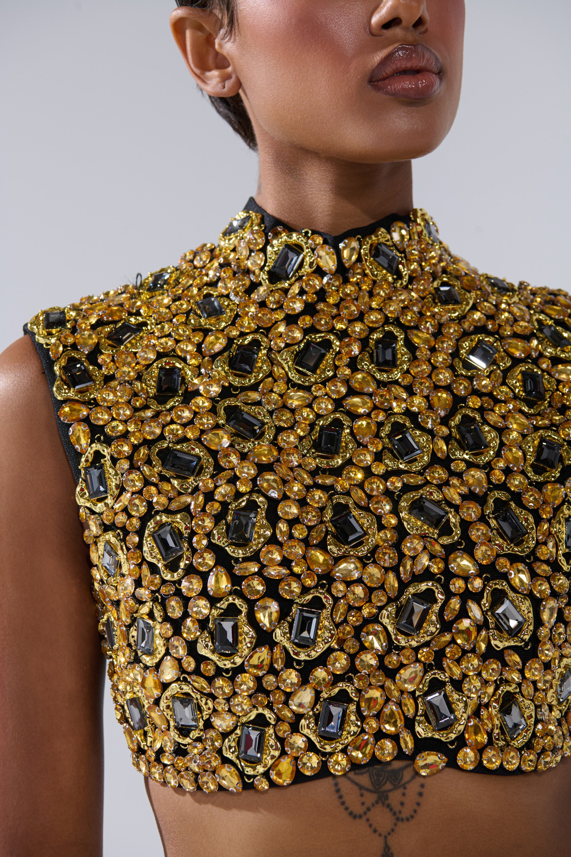 A close-up of a person wearing the GOLDEN GLAM BLOUSE, a sleeveless top adorned with intricate gold and black gemstone embellishments that catch the light, revealing part of a tattoo below the blouse.