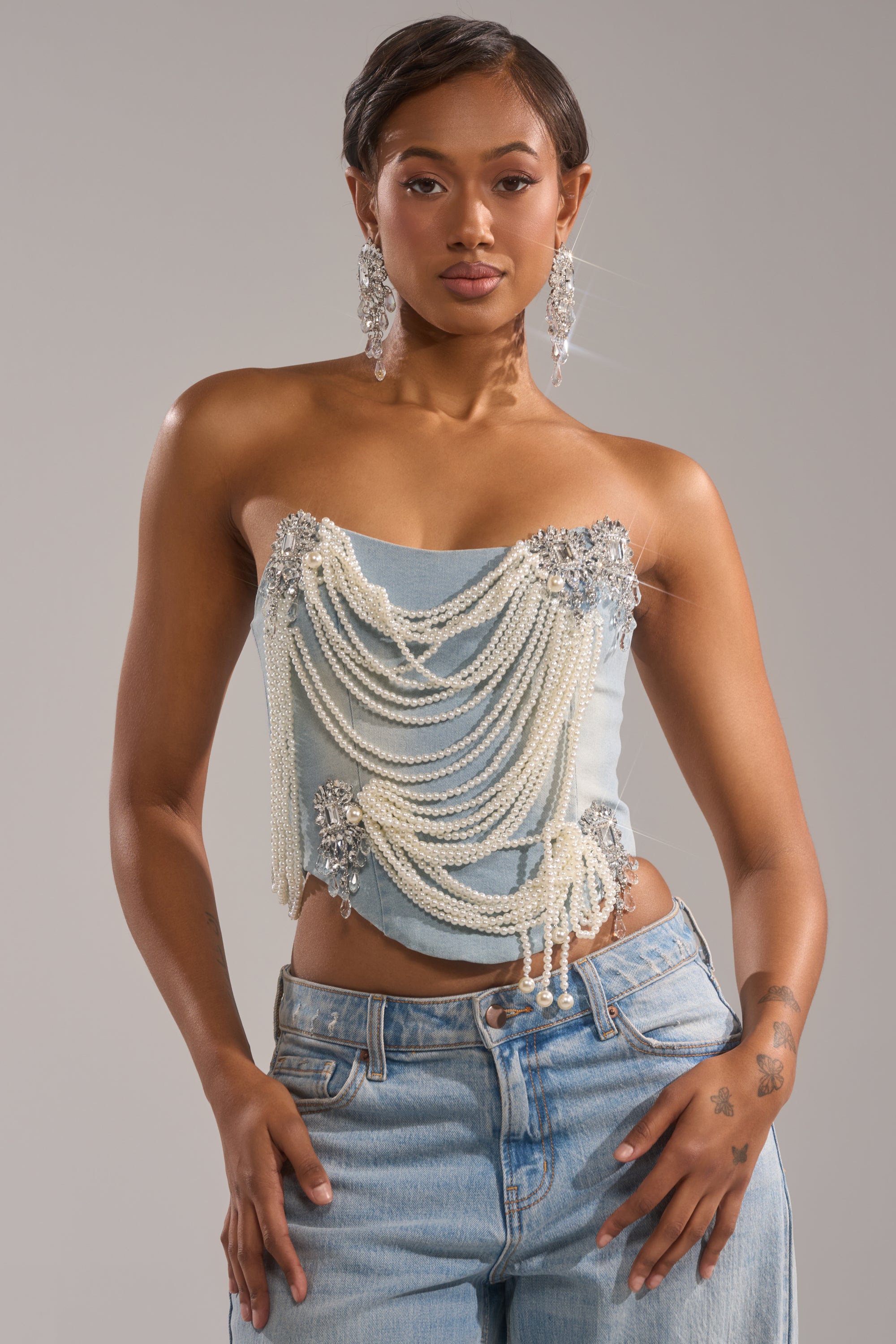 A woman wears the PEARL QUEEN DENIM CORSET adorned with pearl chains and silver accents, paired with light blue jeans, large dangling earrings, and a tattoo on her left forearm.