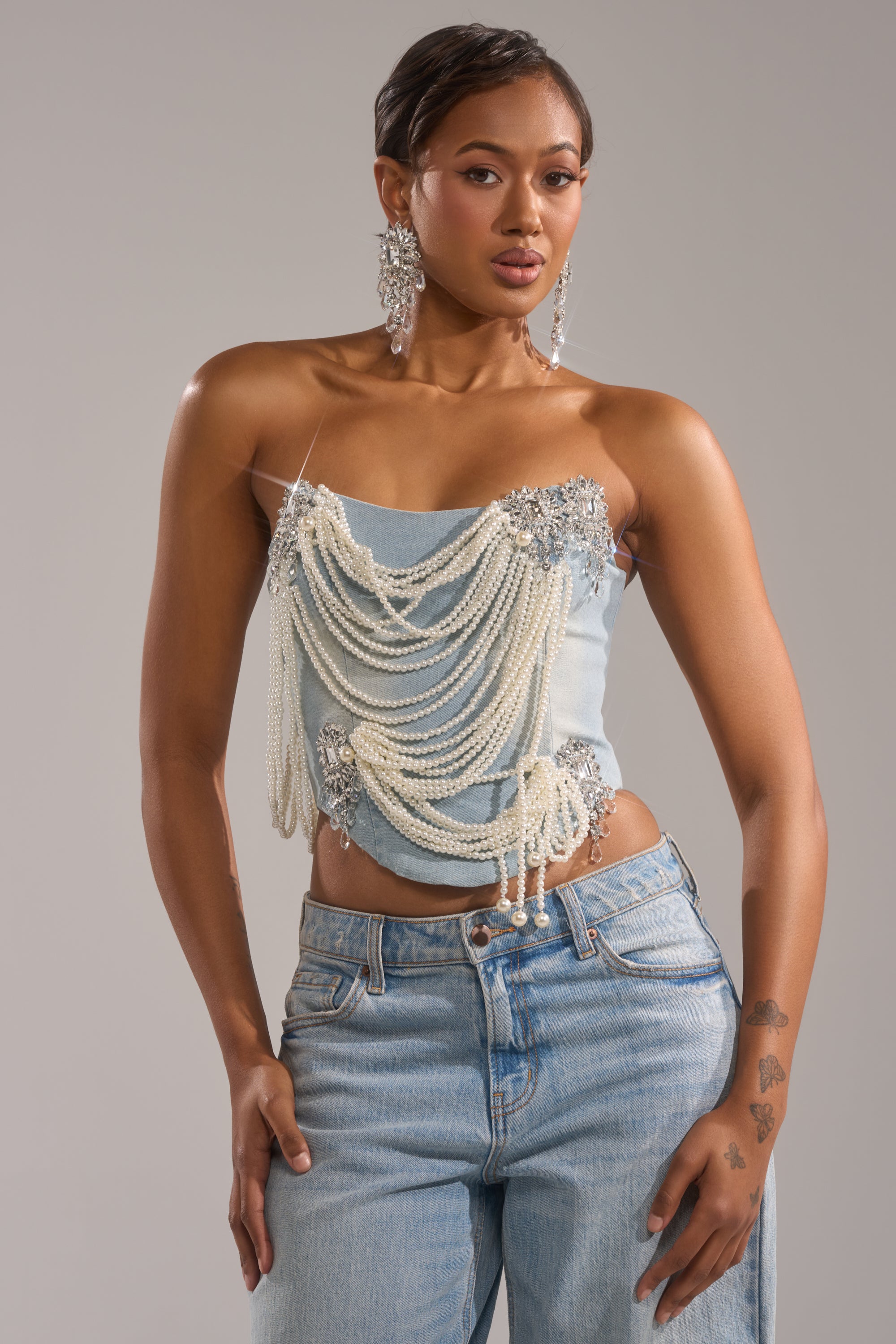 A woman models the PEARL QUEEN DENIM CORSET—adorned with silver embellishments and draped pearl chains—paired with light blue jeans and statement earrings, posing against a plain gray background.